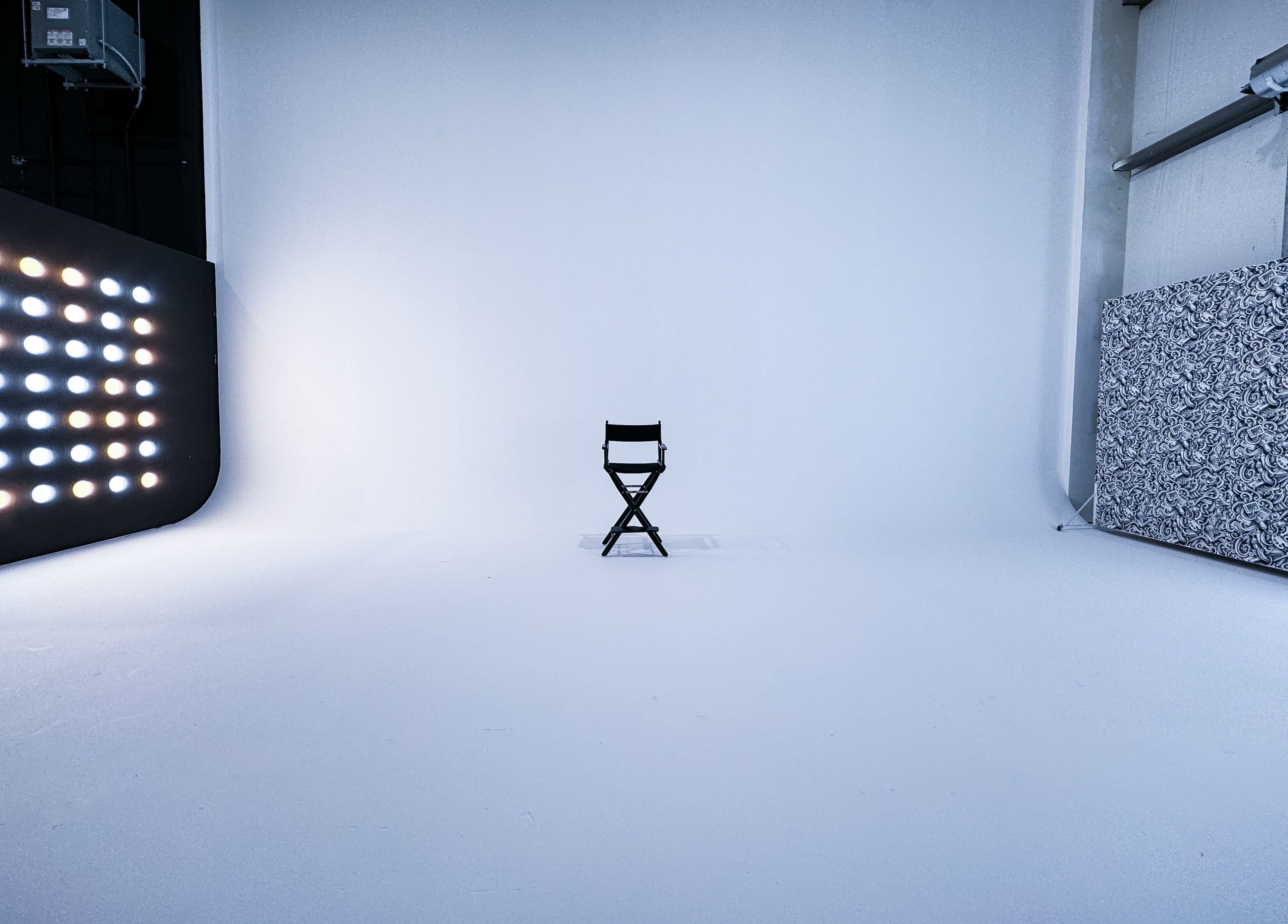 A black director's chair stands alone in a spacious, empty studio with white walls and a lit backdrop on one side.