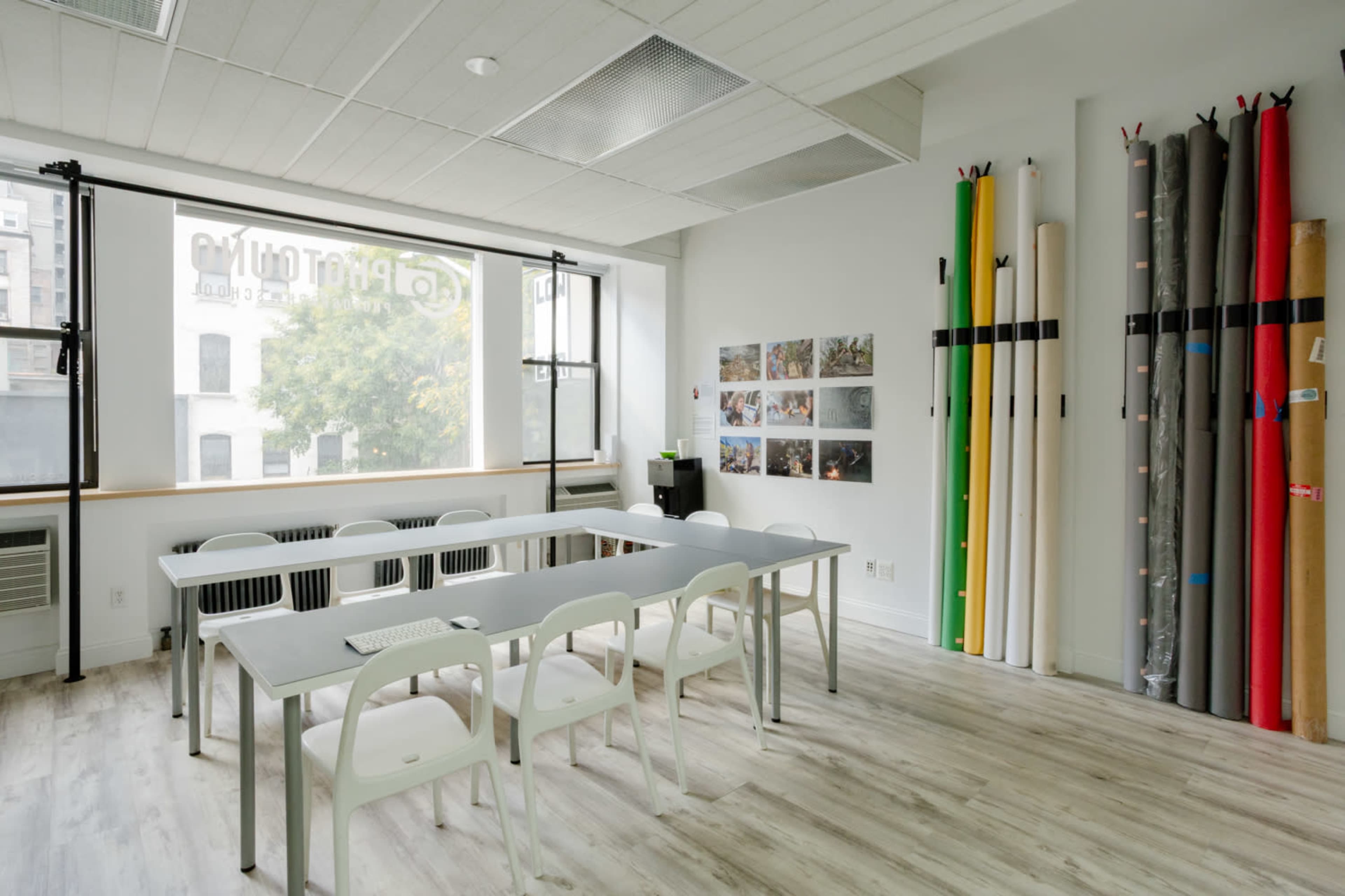 The image shows a bright, modern classroom with a long table and chairs, large windows letting in natural light, and colorful rolls of paper against the wall.