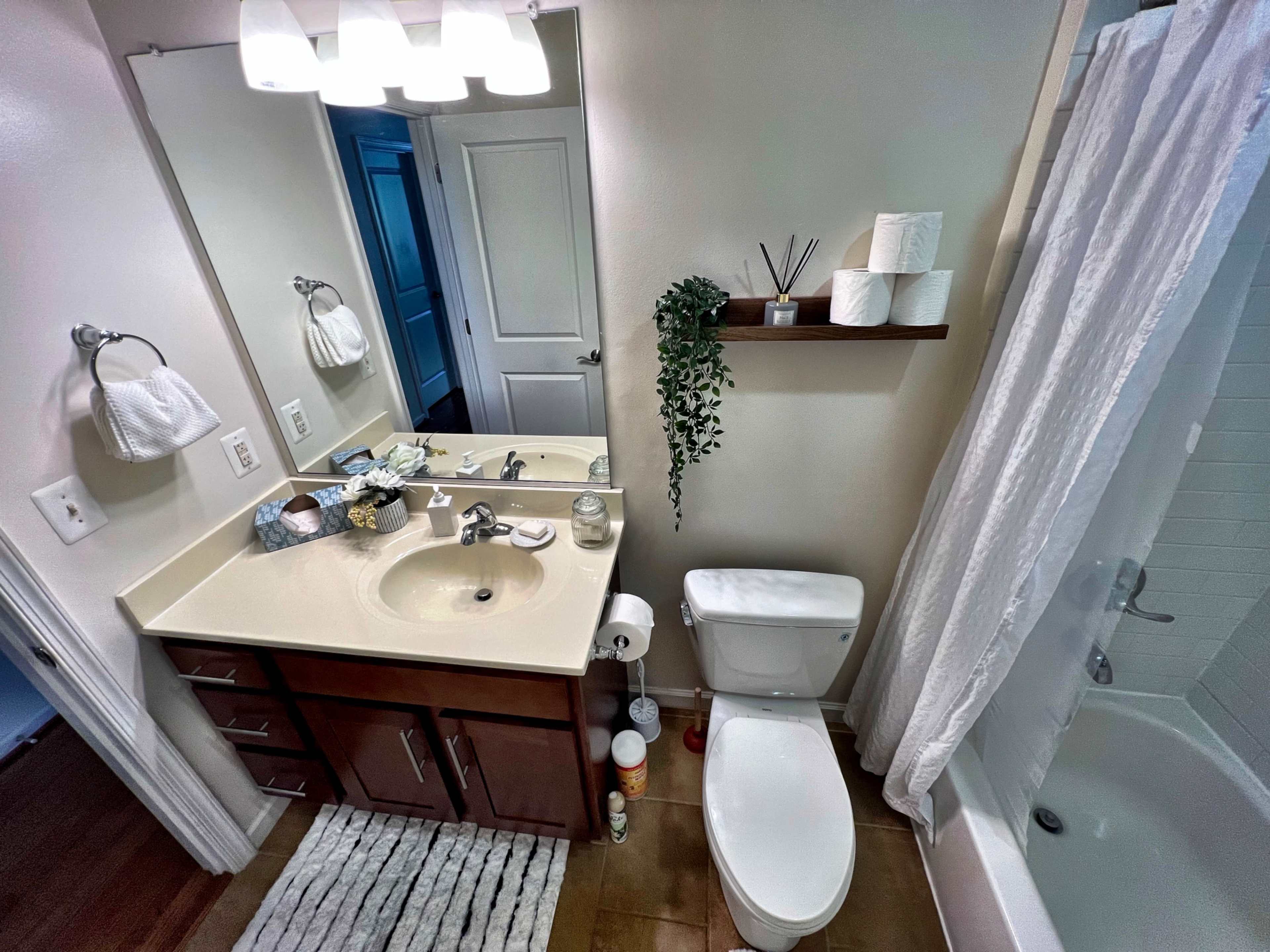 The image shows a bathroom featuring a sink with a mirror, a toilet, and a bathtub with a shower, along with decorative items on a shelf.