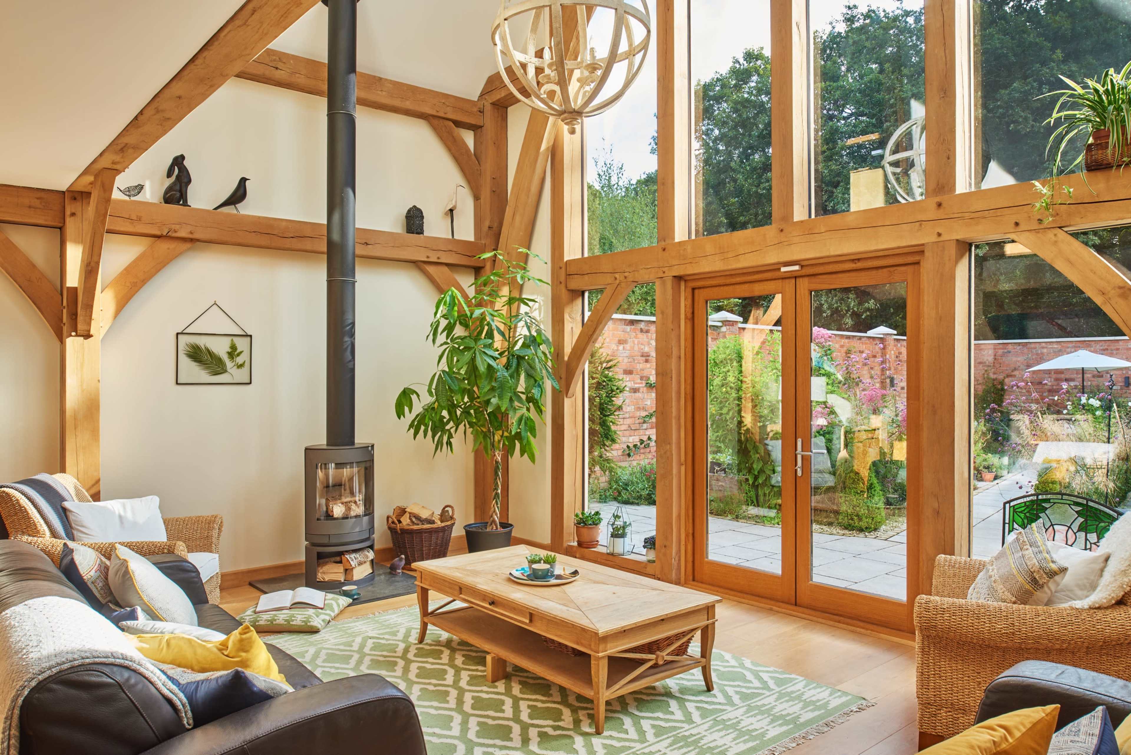 The scene features a bright living room with wooden beams, a wood-burning stove, a large glass door opening to a garden, and a table in the center surrounded by various seating.