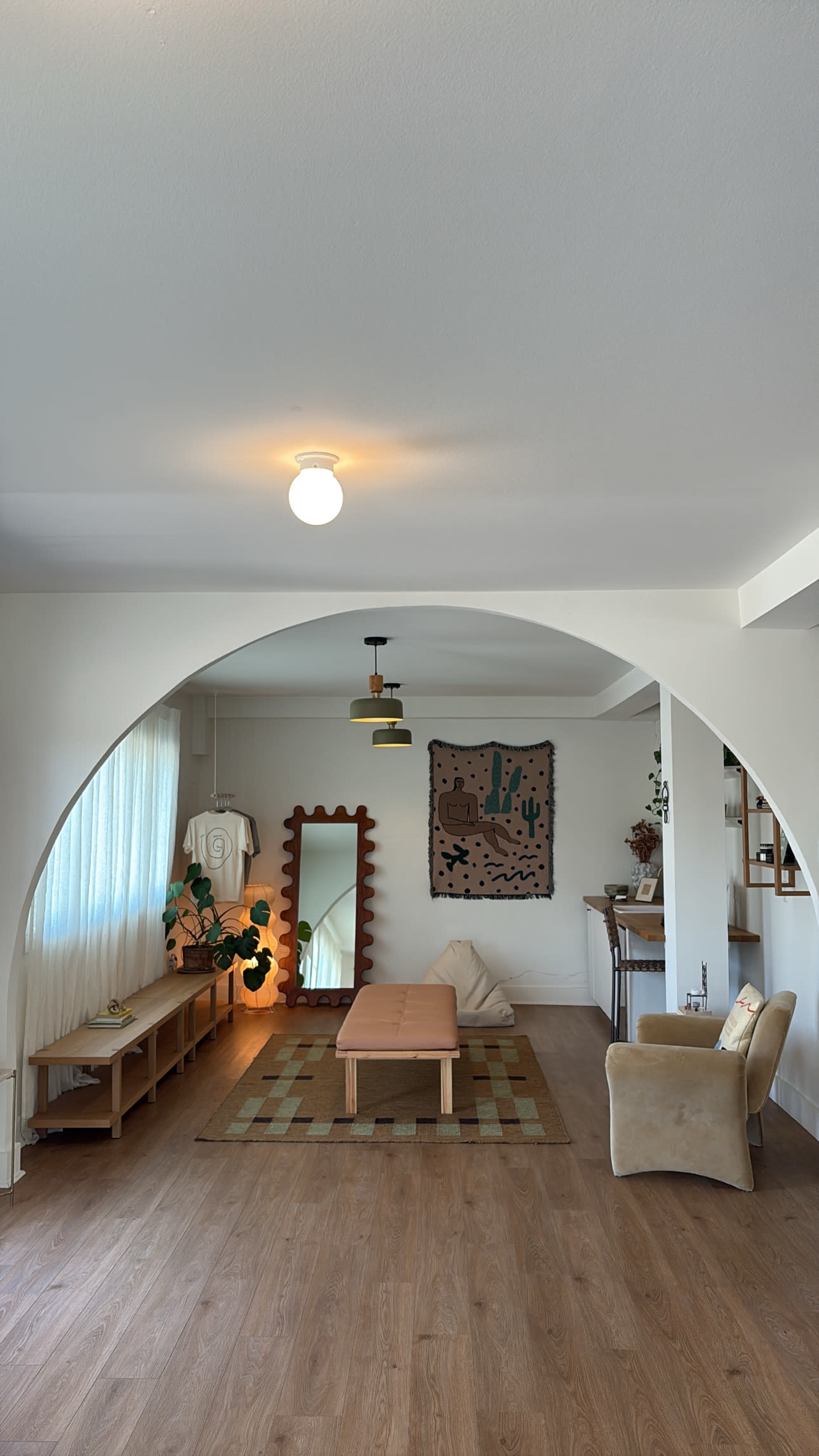 The image shows a stylishly decorated living room featuring an arched doorway, a low coffee table, a large mirror, and various plants.