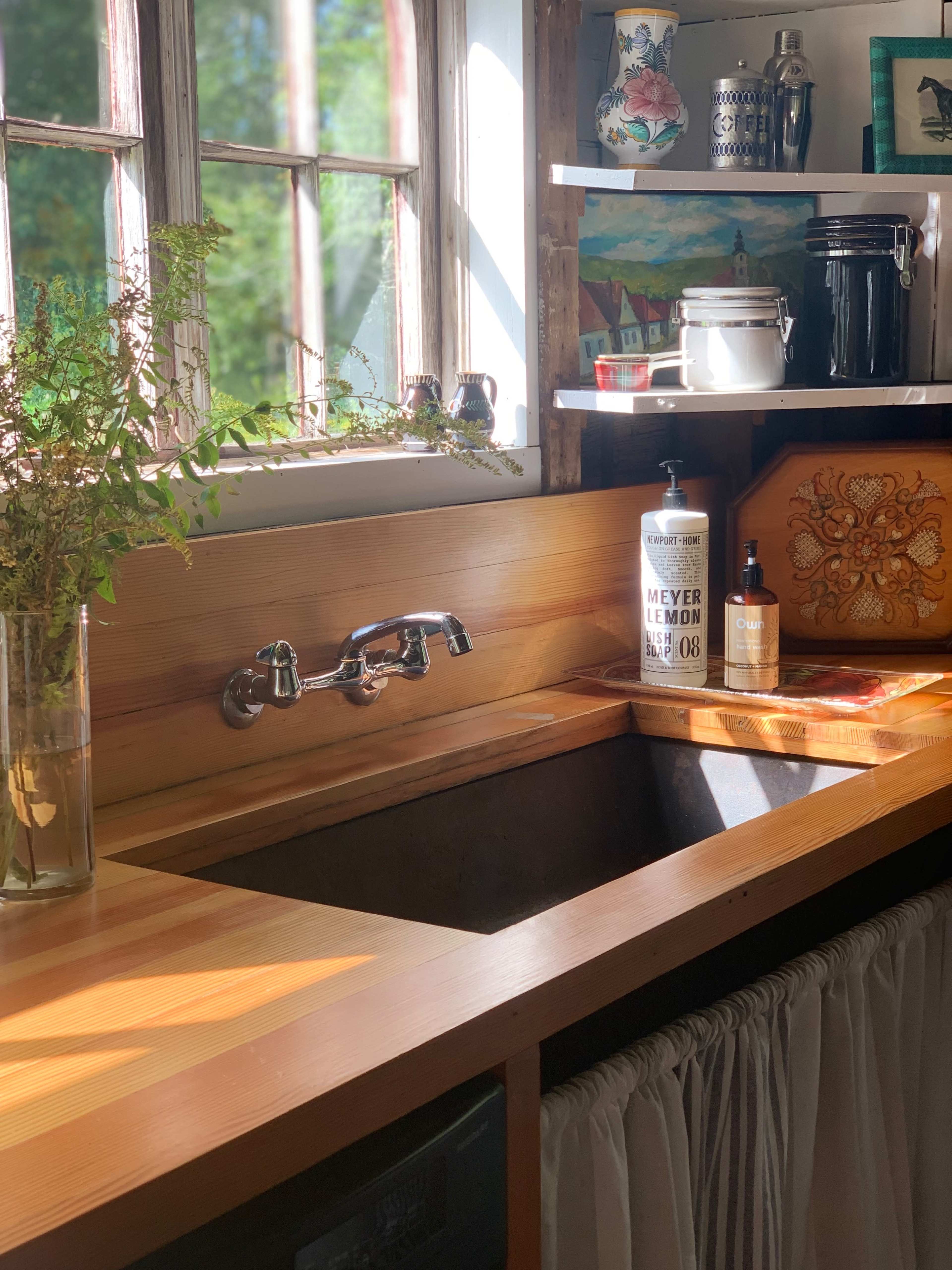 The image shows a wooden kitchen sink area with a metal faucet, a bottle of Meyer lemon dish soap, and a decorative wooden box, all illuminated by natural light coming through a window.