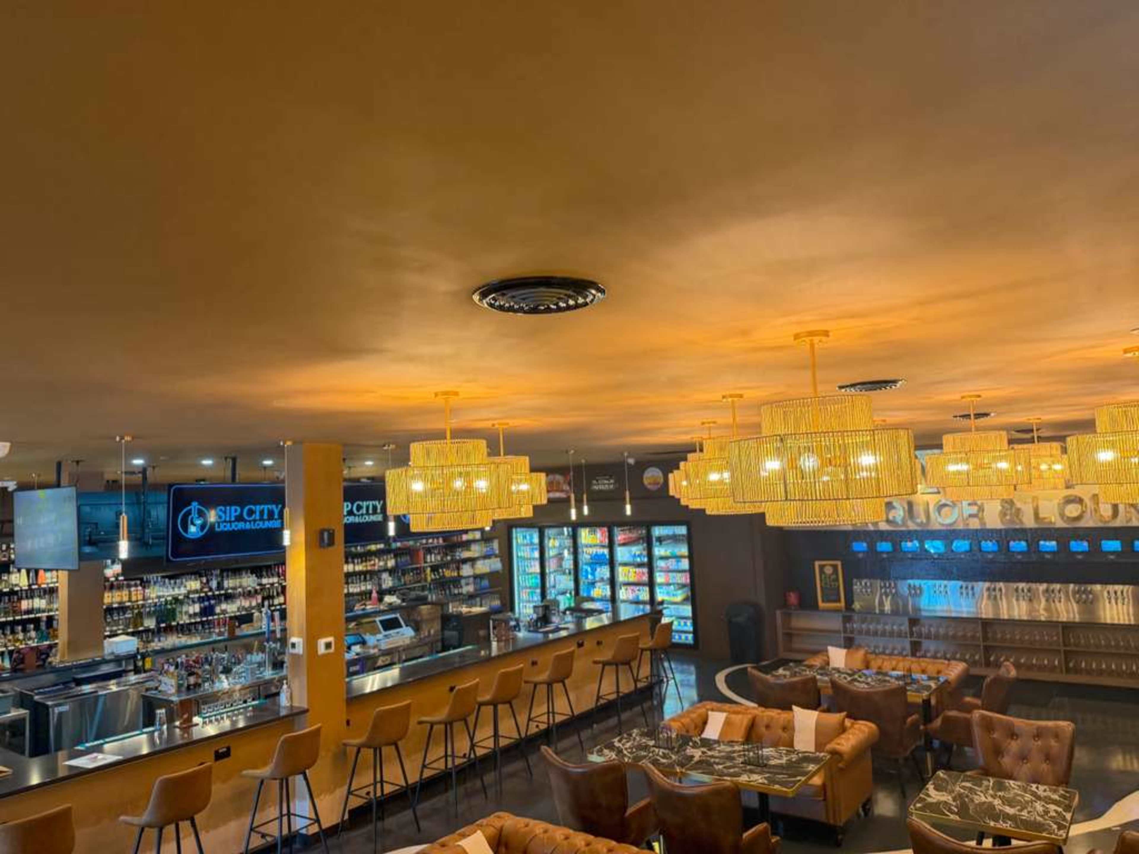 The image shows a modern bar area with a long counter, hanging chandeliers, and a selection of drinks displayed in a refrigerated section.
