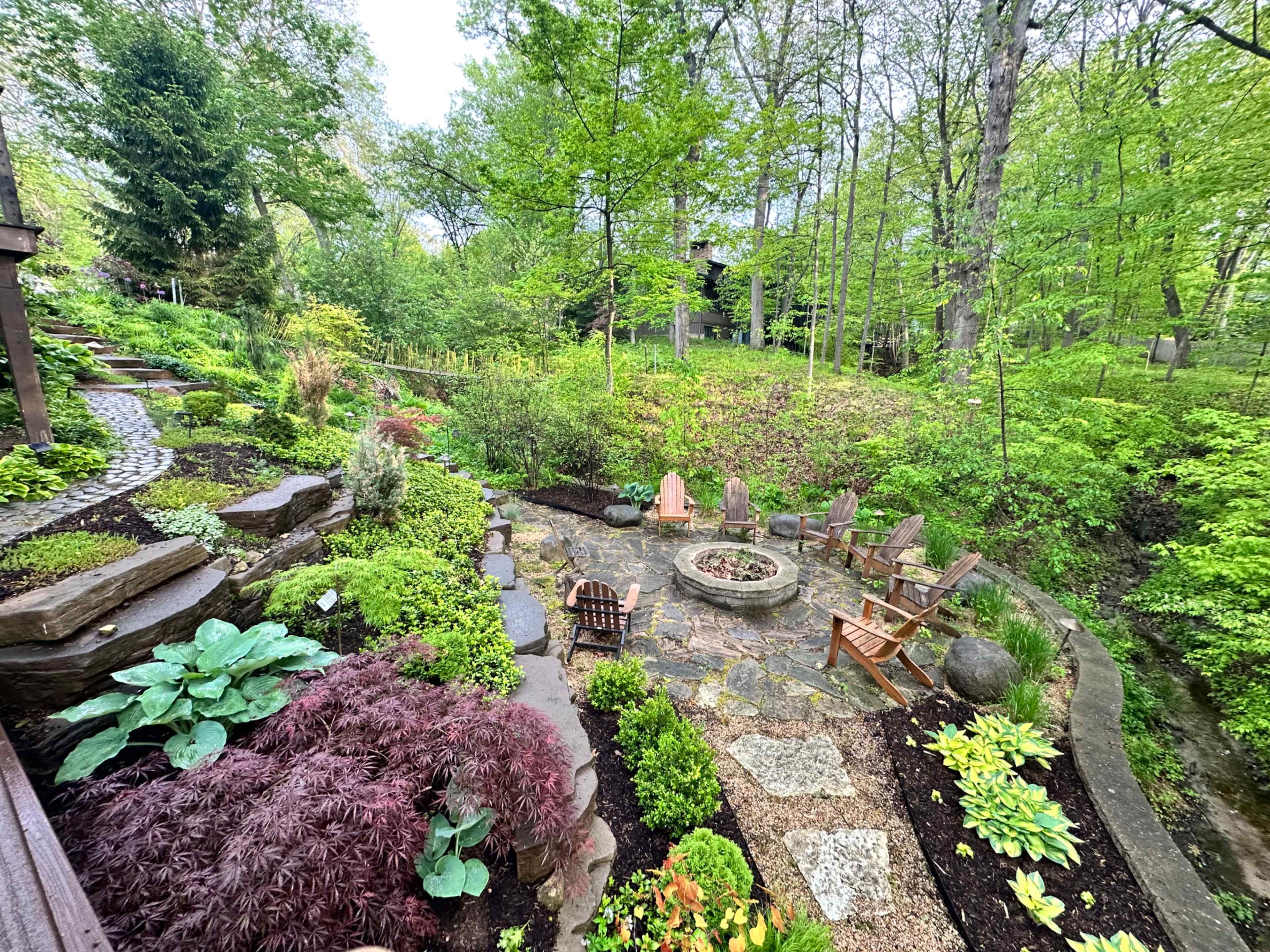 The image shows a landscaped garden with various plants surrounding a circular stone fire pit and several wooden chairs.