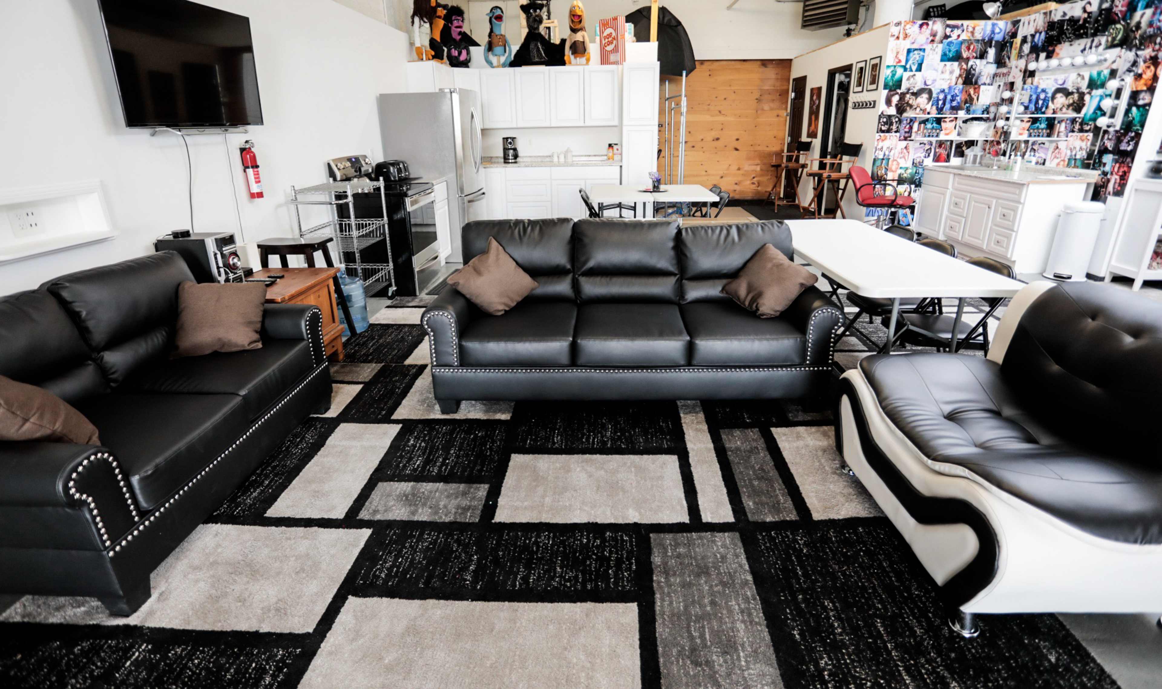 The image shows a modern living space with black leather sofas, a coffee table, and a kitchen area in the background featuring a refrigerator and white cabinetry.