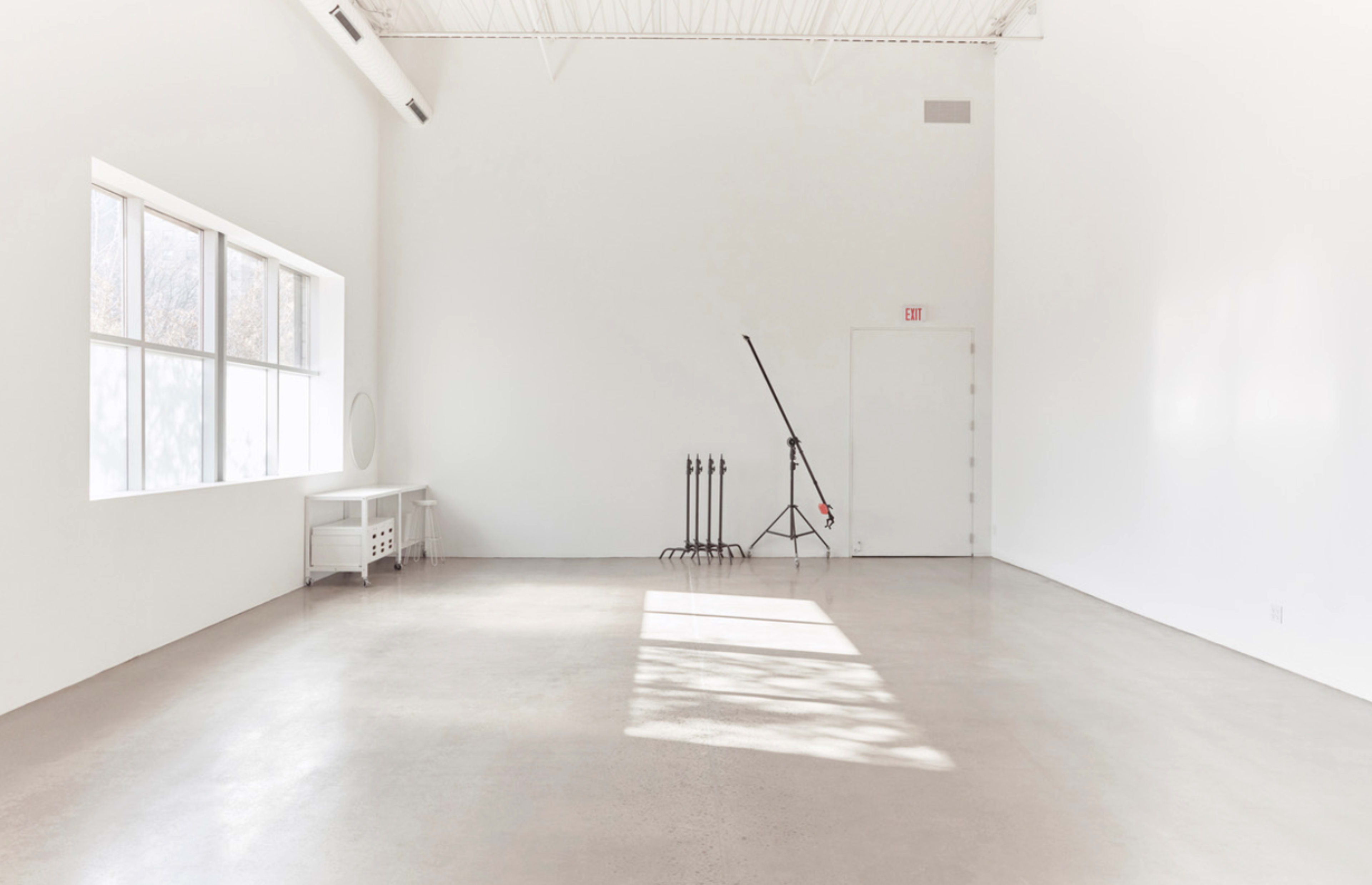 A bright, empty room with white walls, a large window, a door, and a lighting stand in the corner.