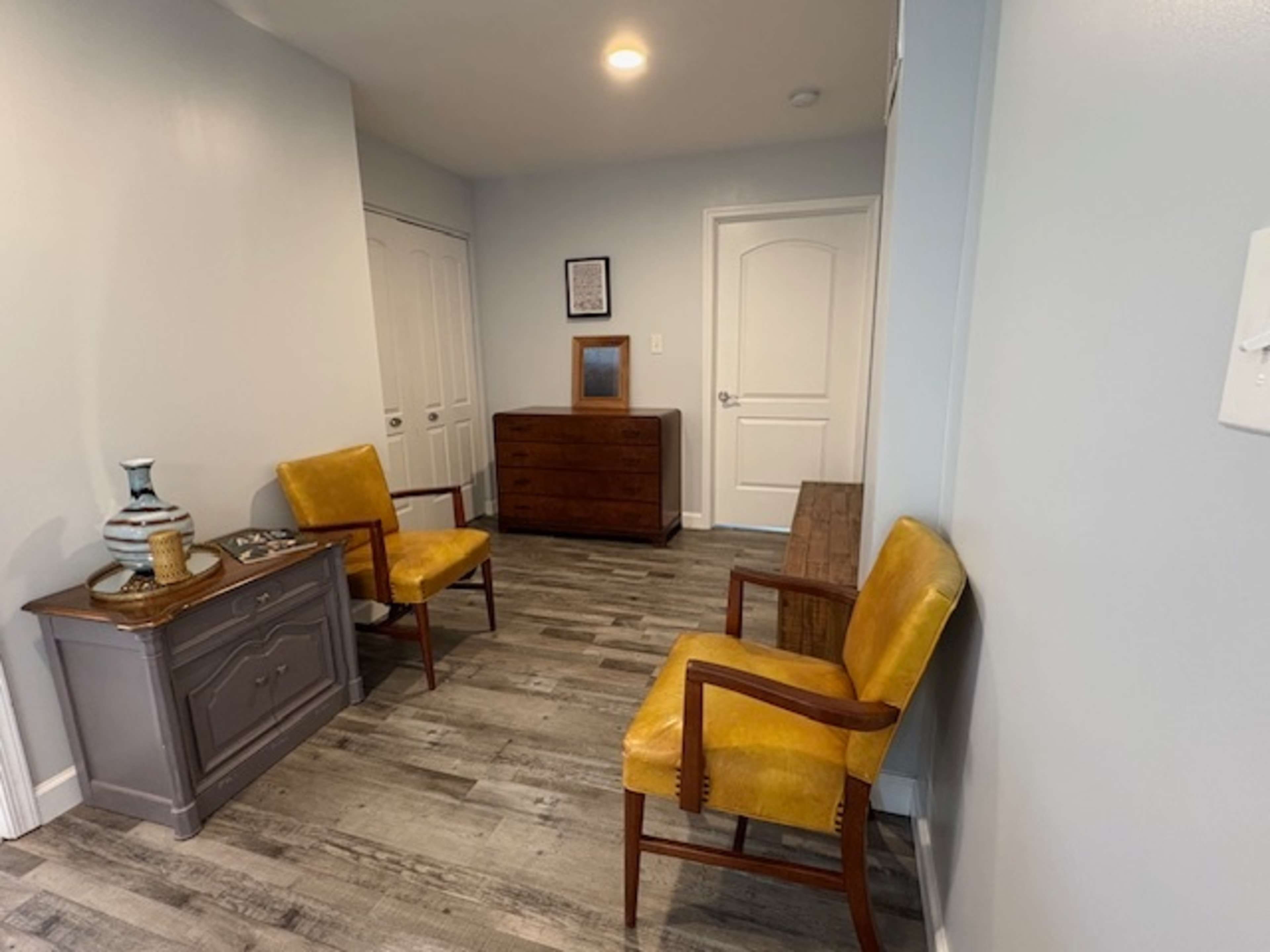 The image shows a small, well-lit hallway with two yellow chairs, a wooden dresser, and a side table against the walls.