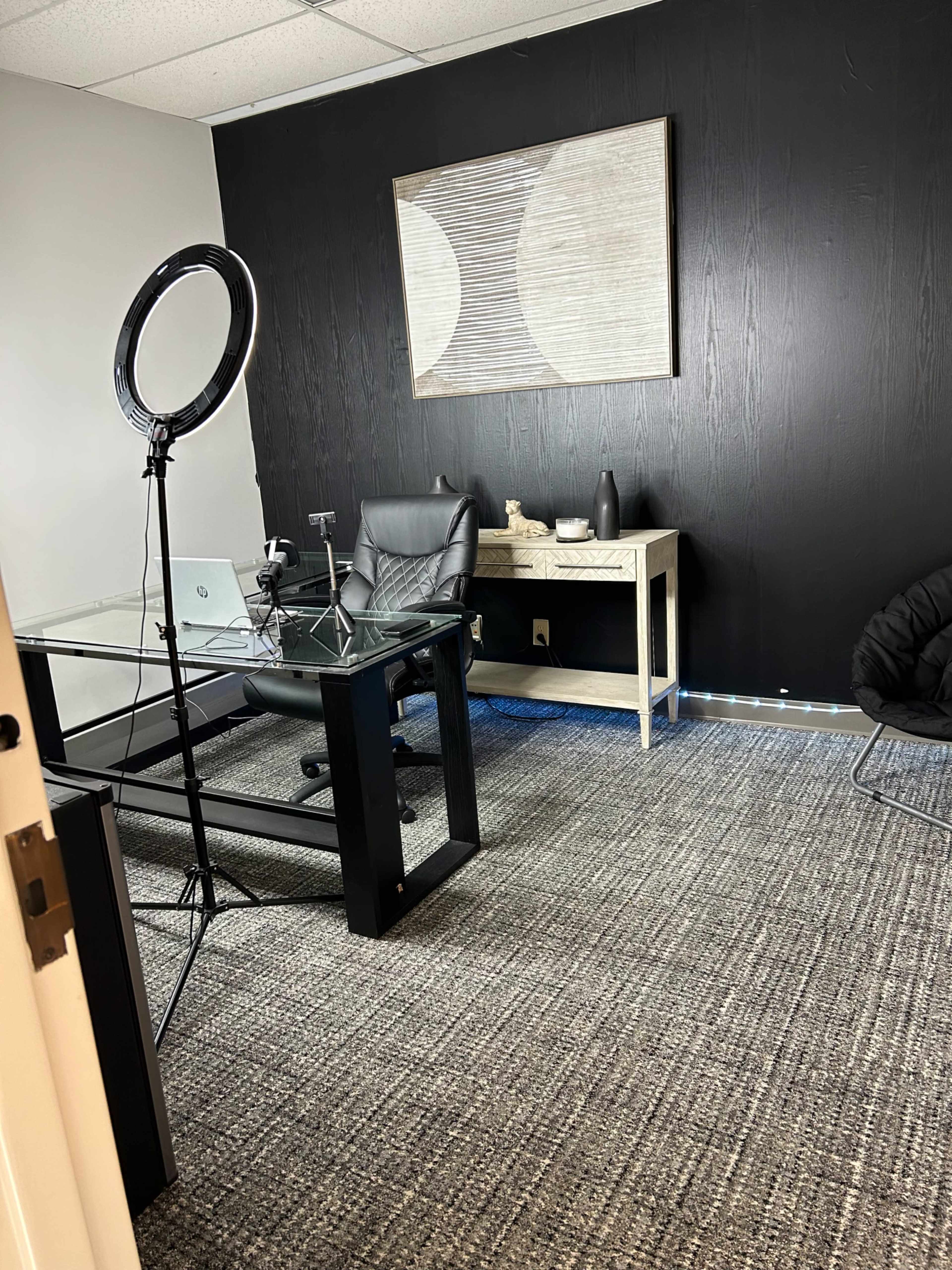A dimly lit office space featuring a glass desk, a black chair, a decorative wall art piece, and a ring light setup on a tripod.