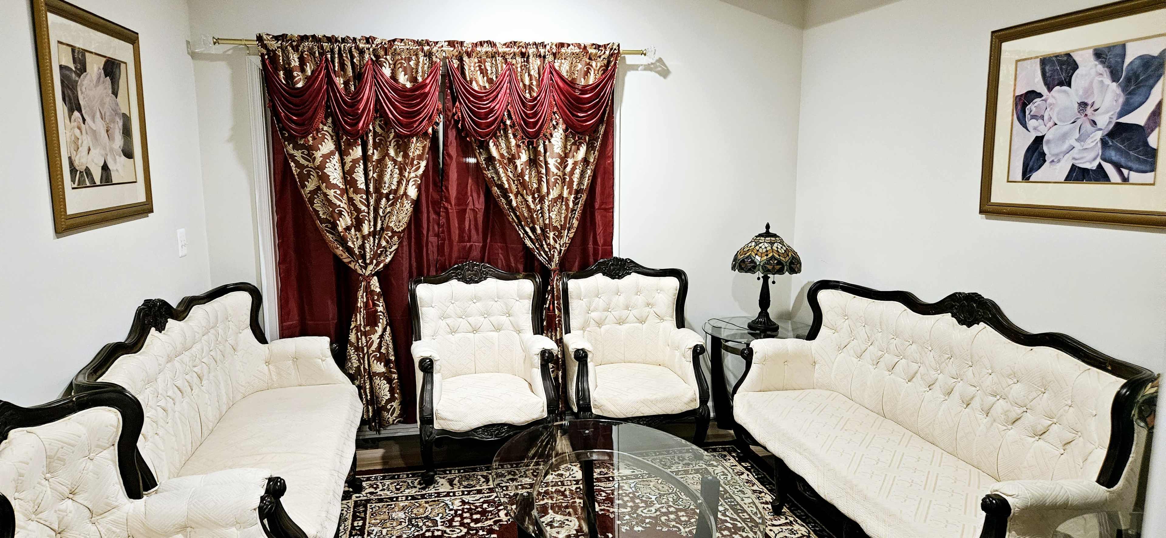 The image depicts a living room with ornate cream-colored upholstered sofas arranged around a glass coffee table, complemented by rich red curtains and floral artwork on the walls.