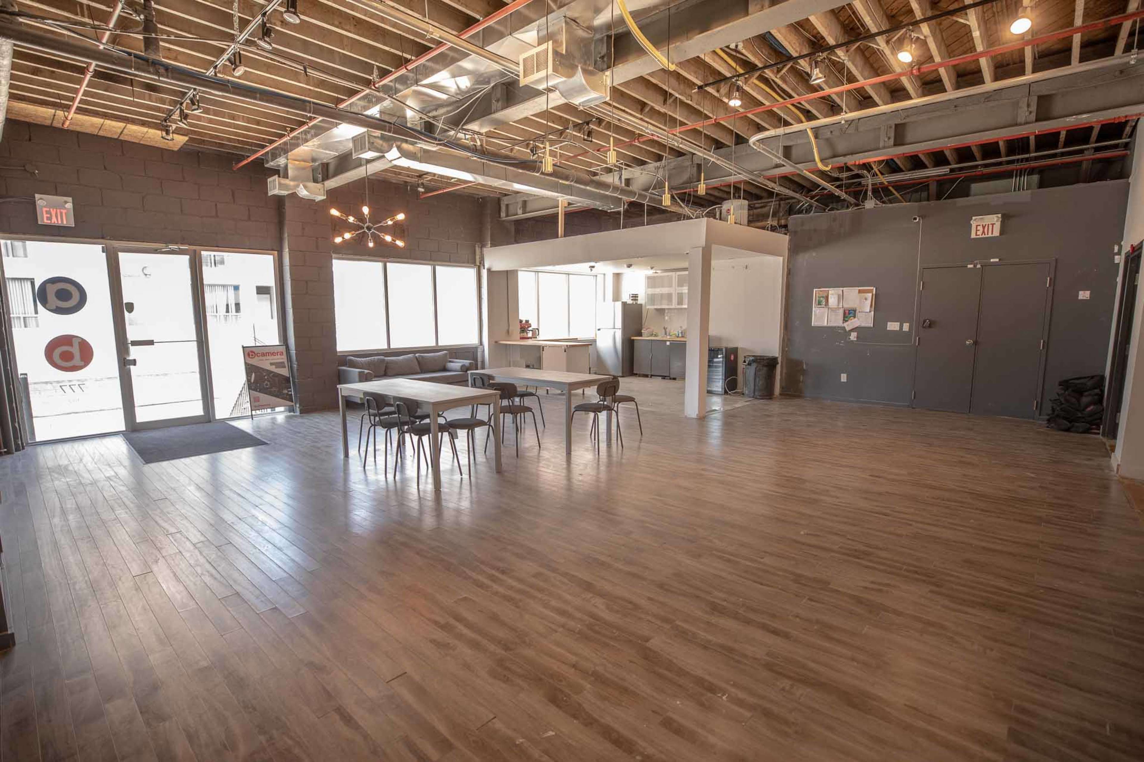 The image shows a spacious, modern interior of a building with wooden floors, overhead industrial lighting, and a seating area that includes a dining table and a kitchenette.