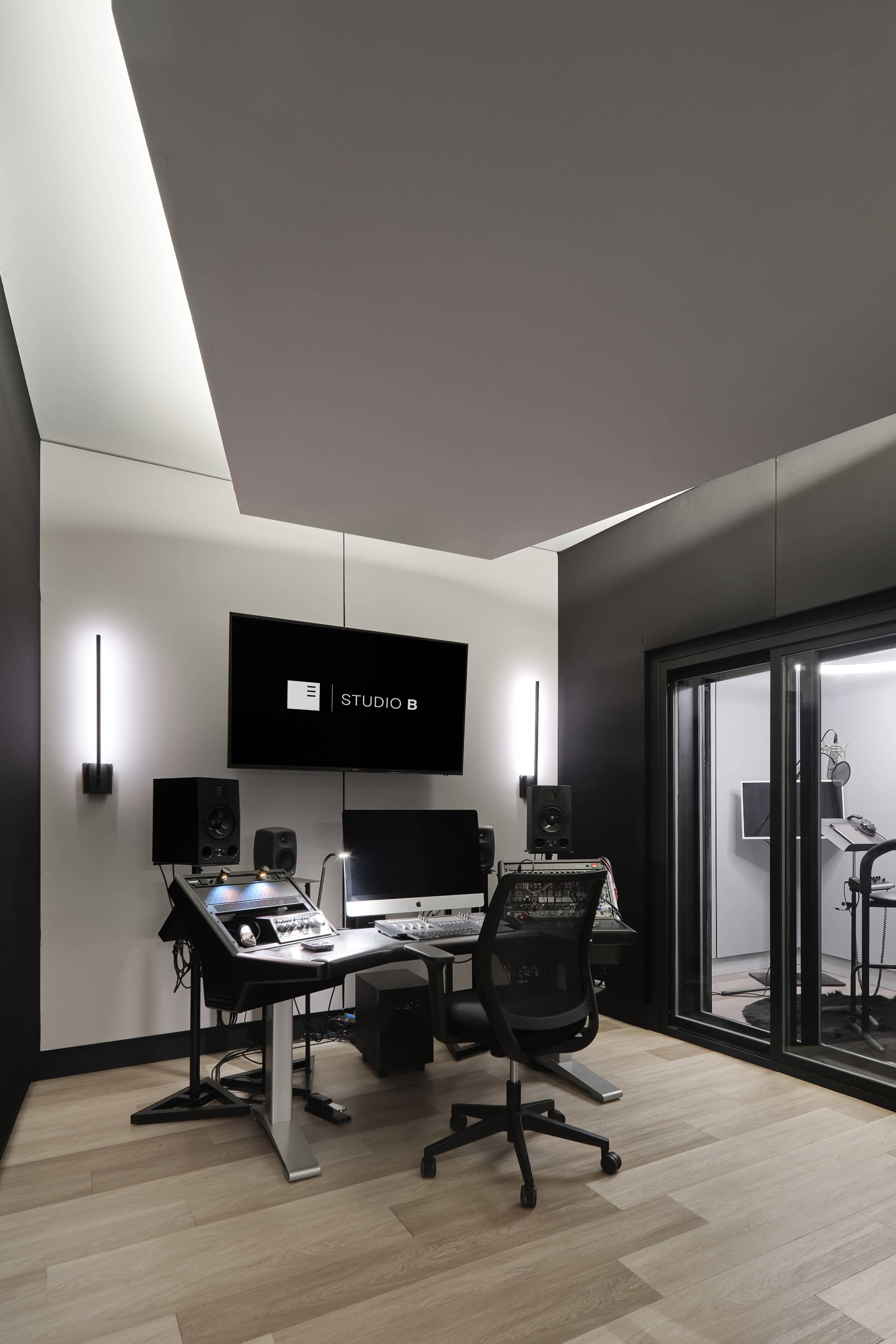 The image shows a modern music studio featuring a desk with computer equipment, audio monitors, and a glass-enclosed sound booth.