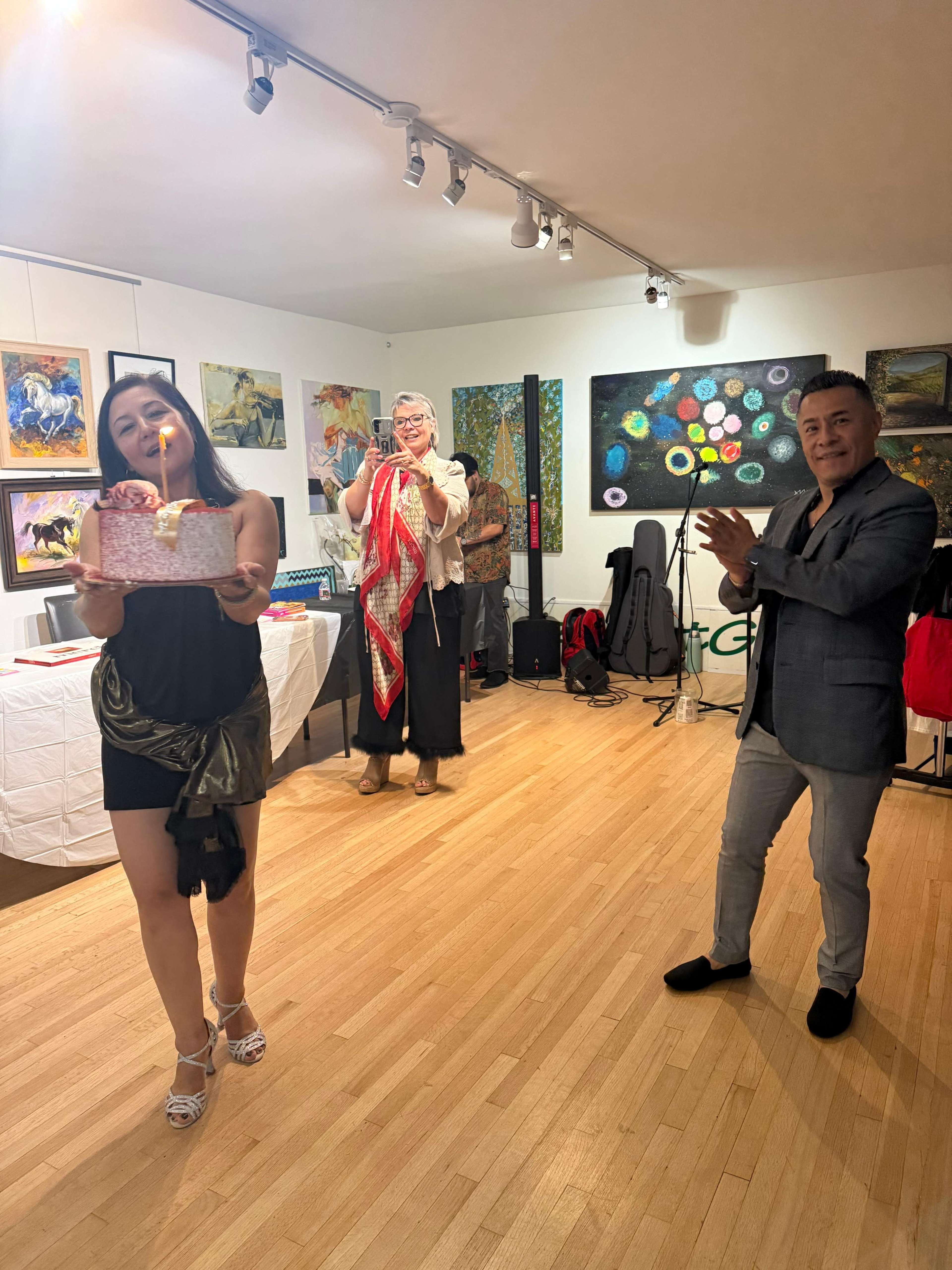 The image depicts a small gallery with three individuals: one woman in a black dress holding a gift, a second woman in a colorful shawl, and a man in a gray suit, all in front of various artworks.