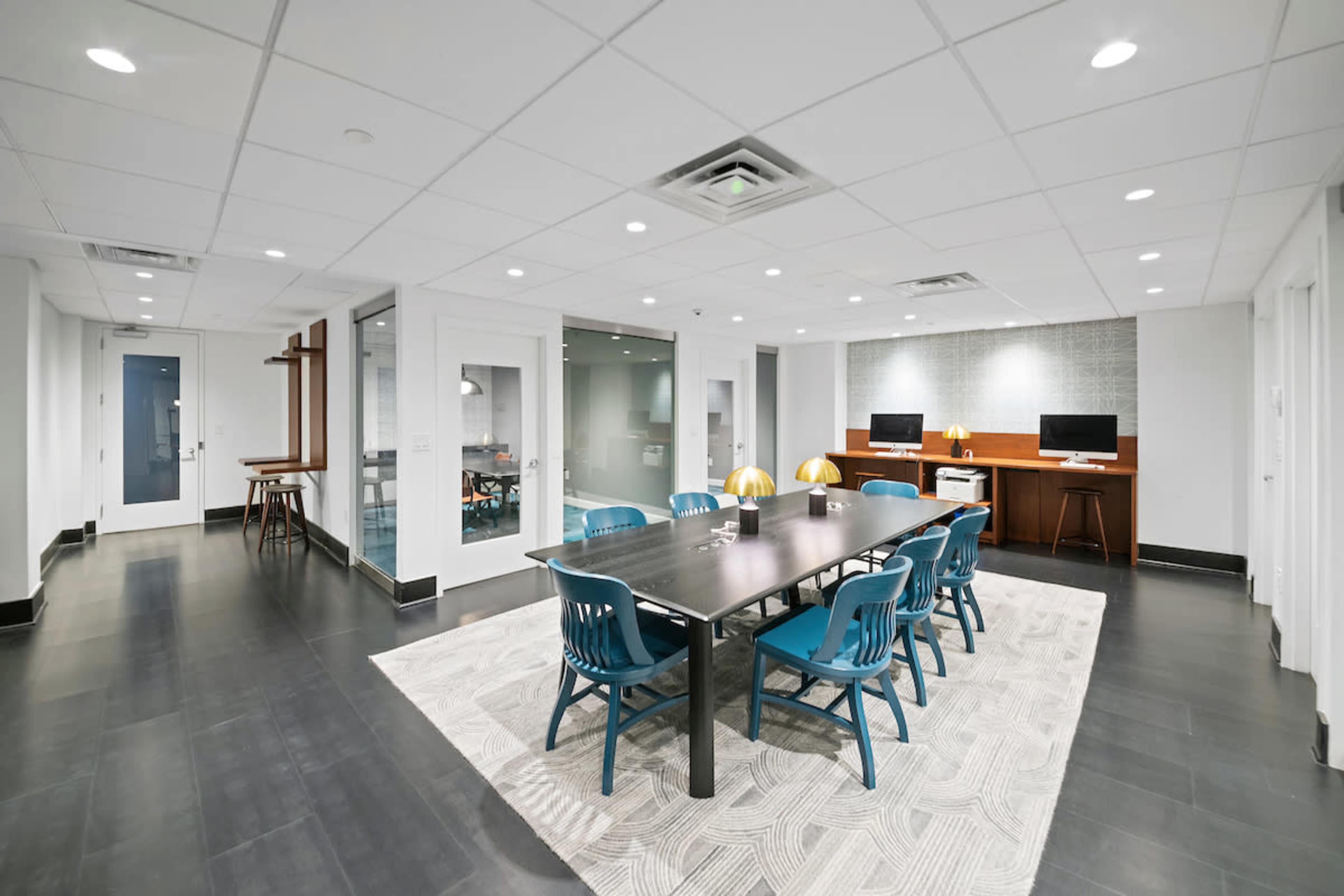 The image shows a modern office space featuring a long table surrounded by blue chairs, with a separate area containing two computers and a decorative lamp.