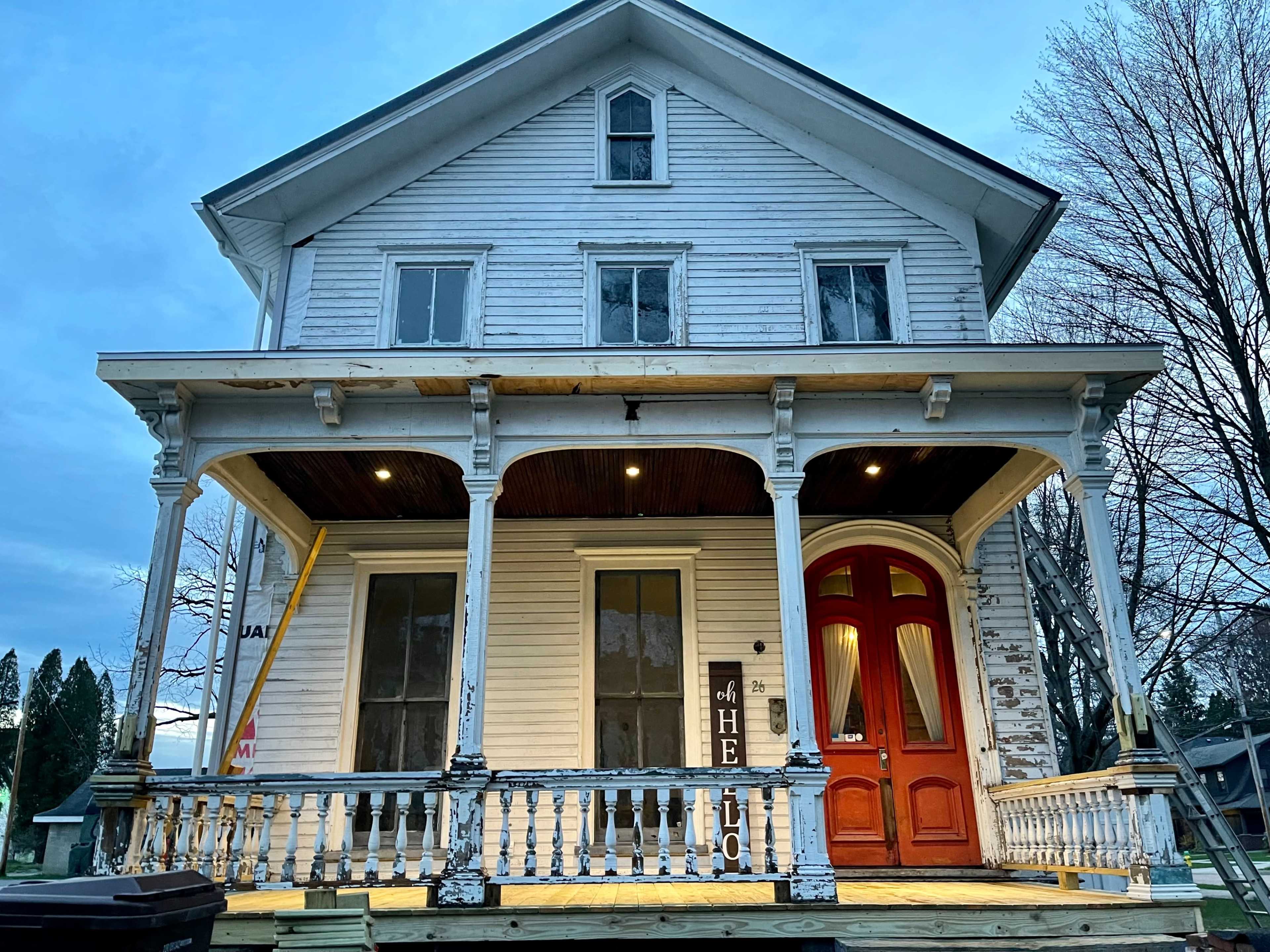 The image shows a large, three-story Victorian-style house with a wraparound porch, featuring white siding and bright red doors.