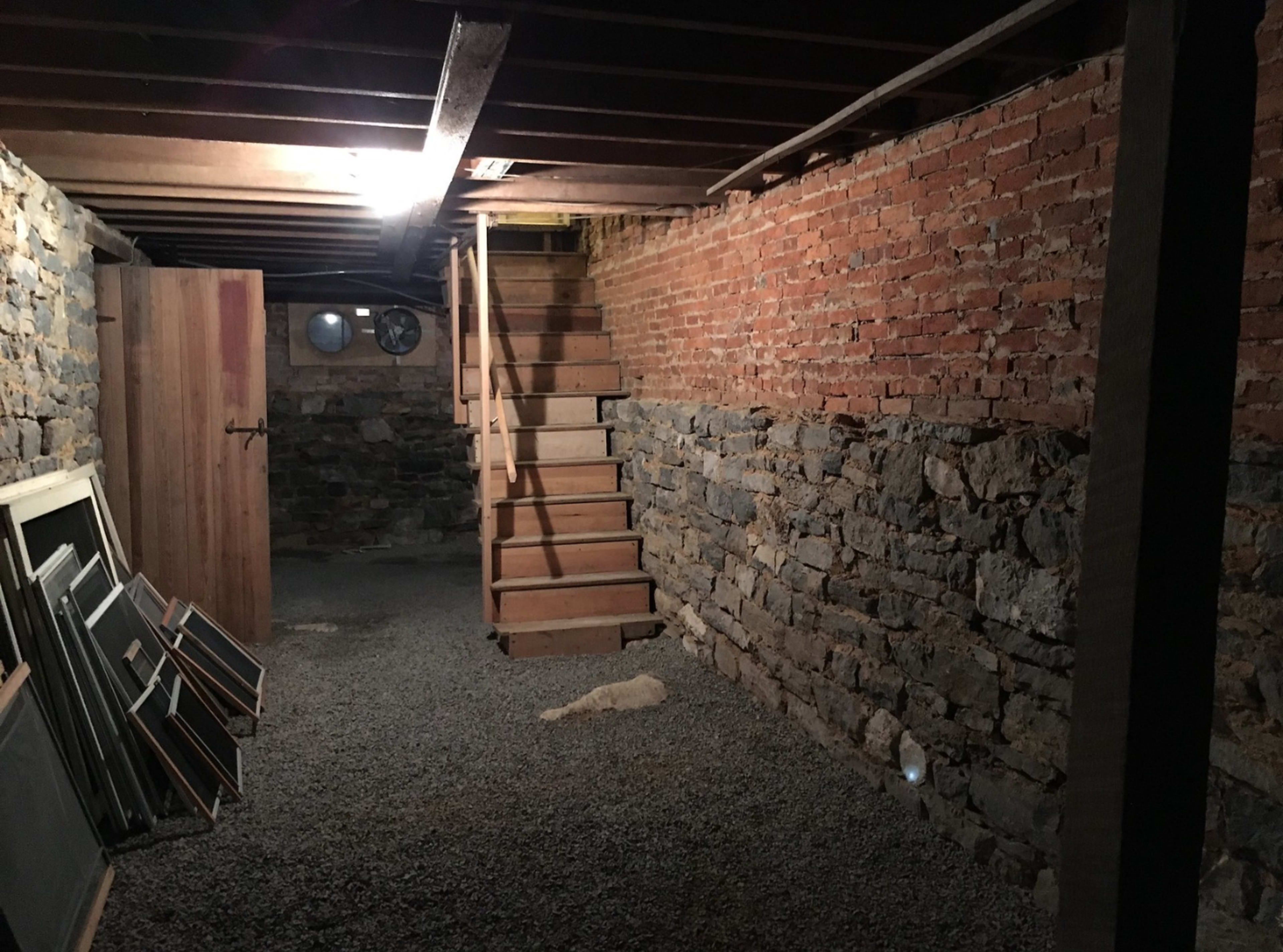 A basement with exposed stone and brick walls, wooden beams, and a staircase leading up, along with stacked frames against one wall.