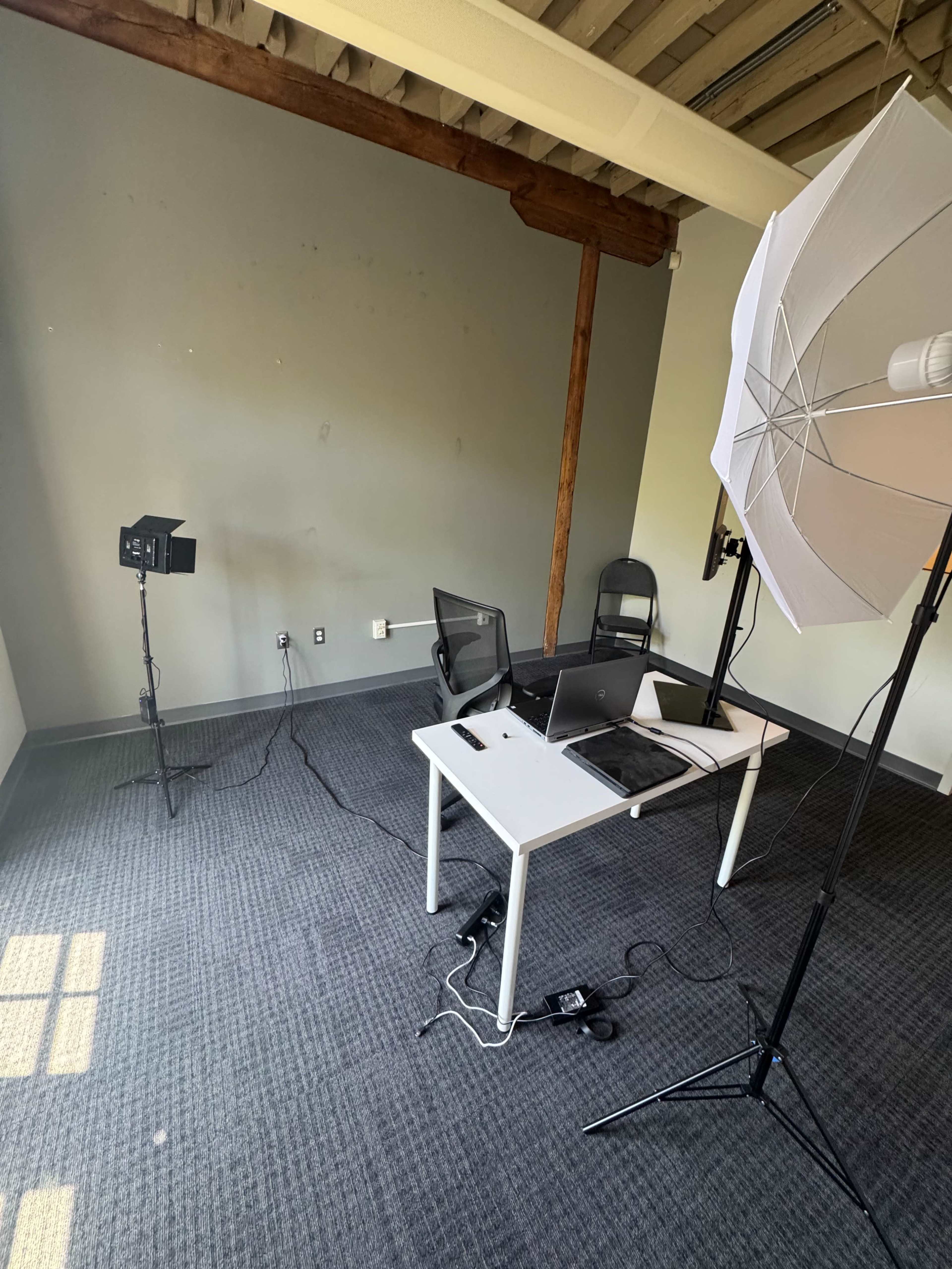 The image shows a sparsely furnished office space with a table, two chairs, and lighting equipment set up for a video shoot.