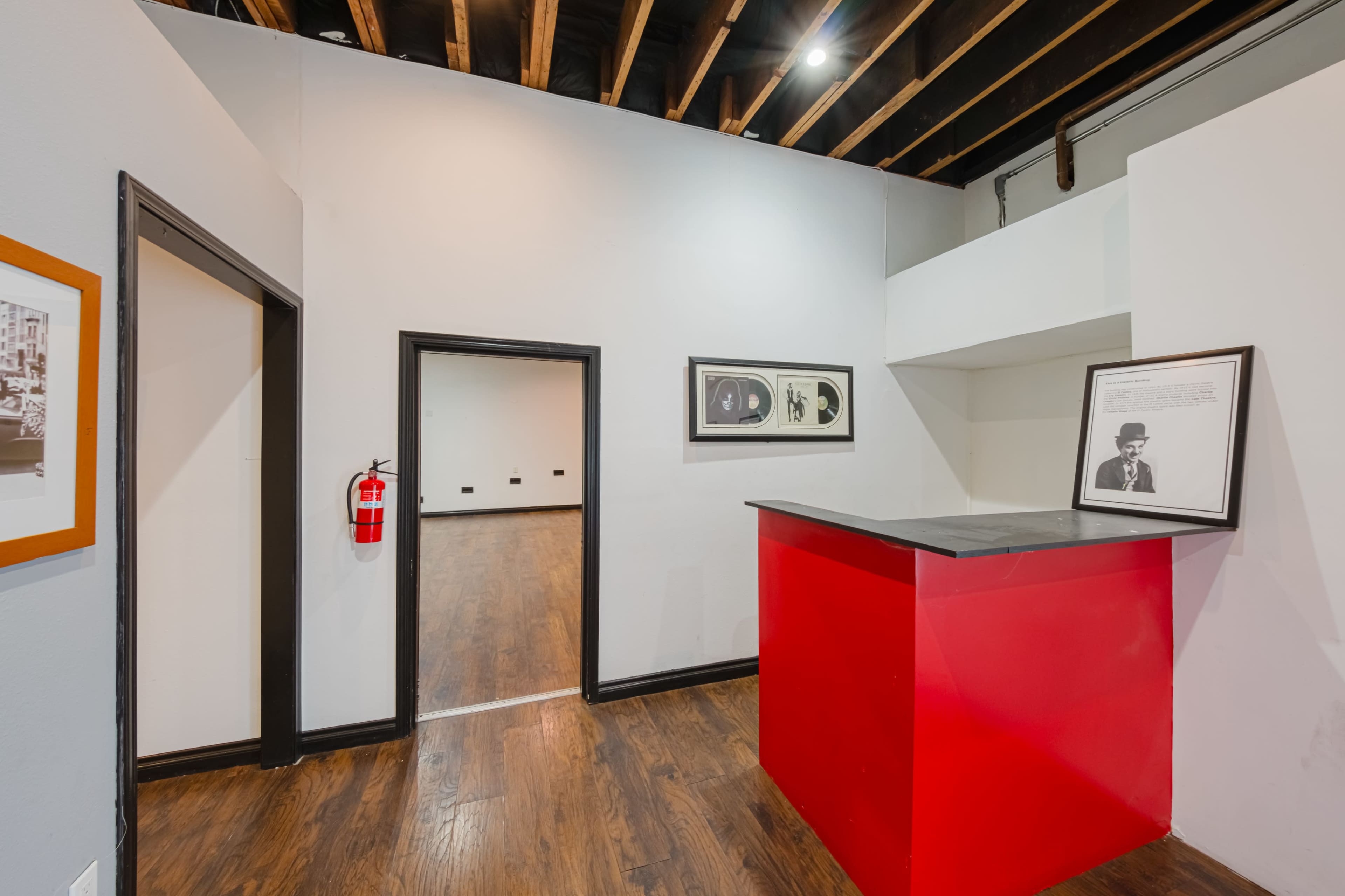 A spacious interior with wooden flooring, a red reception counter on the right, framed photographs on the walls, and an open doorway leading to another room.