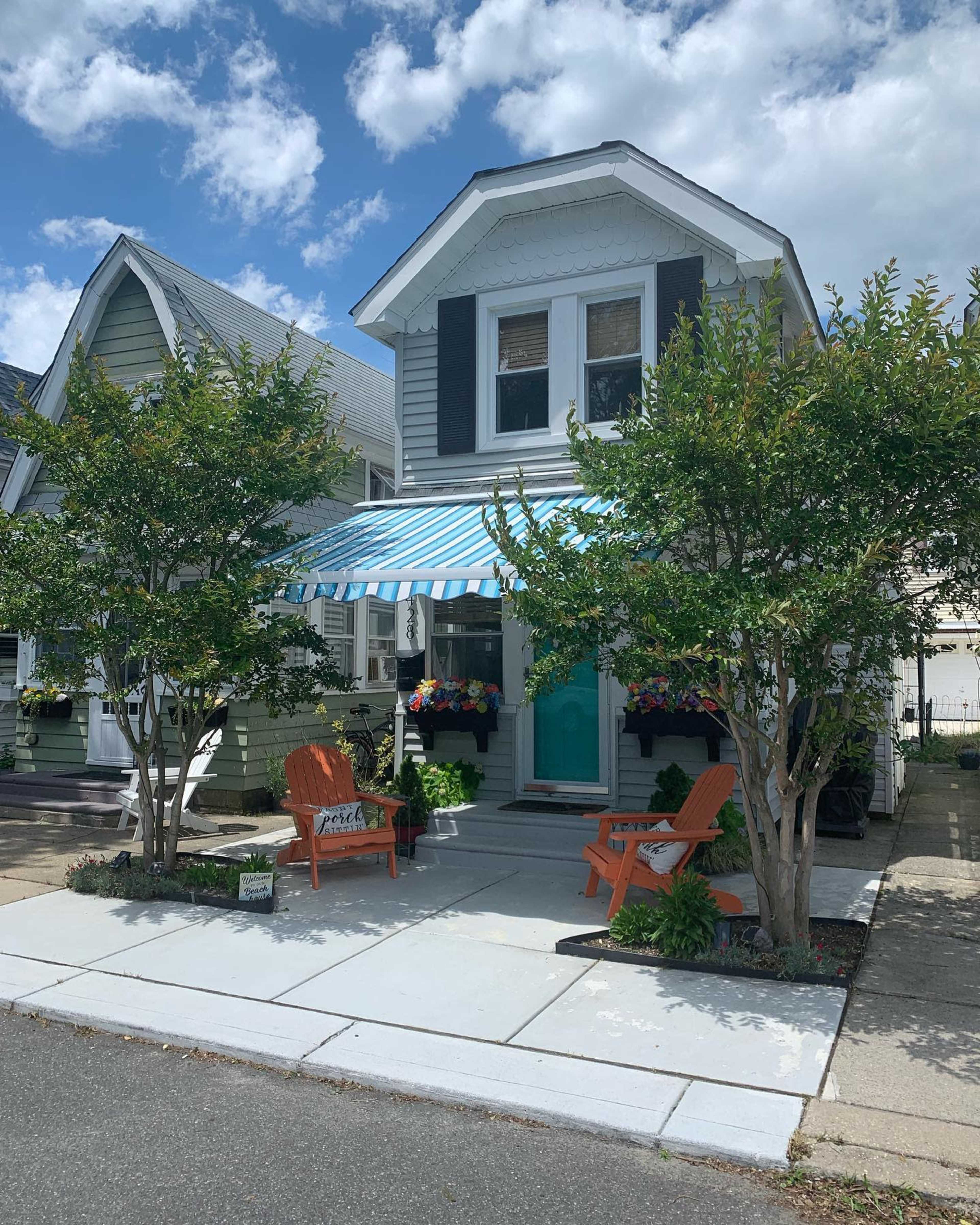 A two-story house with a blue and white striped awning, adorned with potted plants, features a welcoming porch with orange chairs.