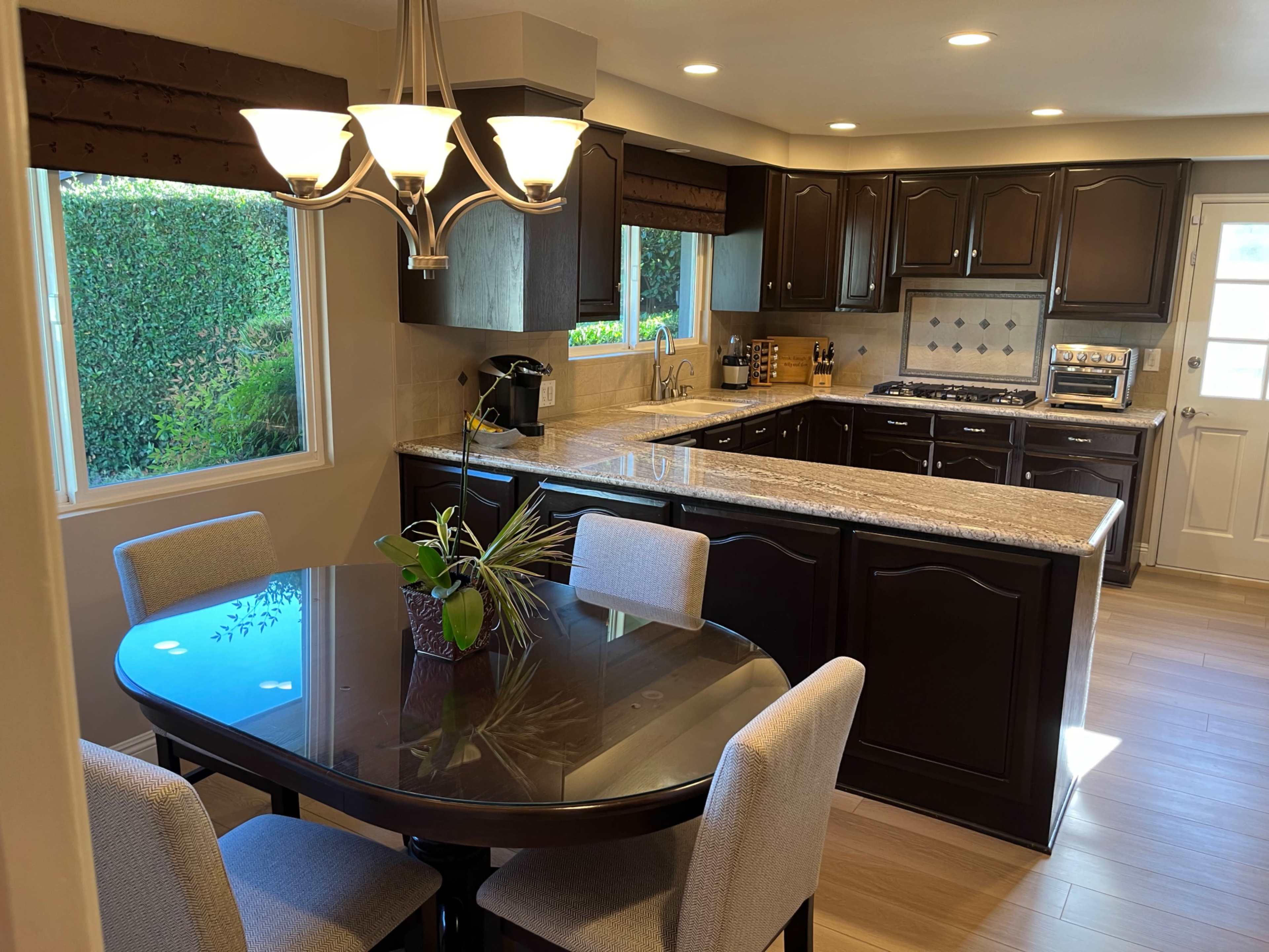 The image depicts a modern kitchen with dark cabinetry, a granite countertop, and a dining area featuring a round table and upholstered chairs.