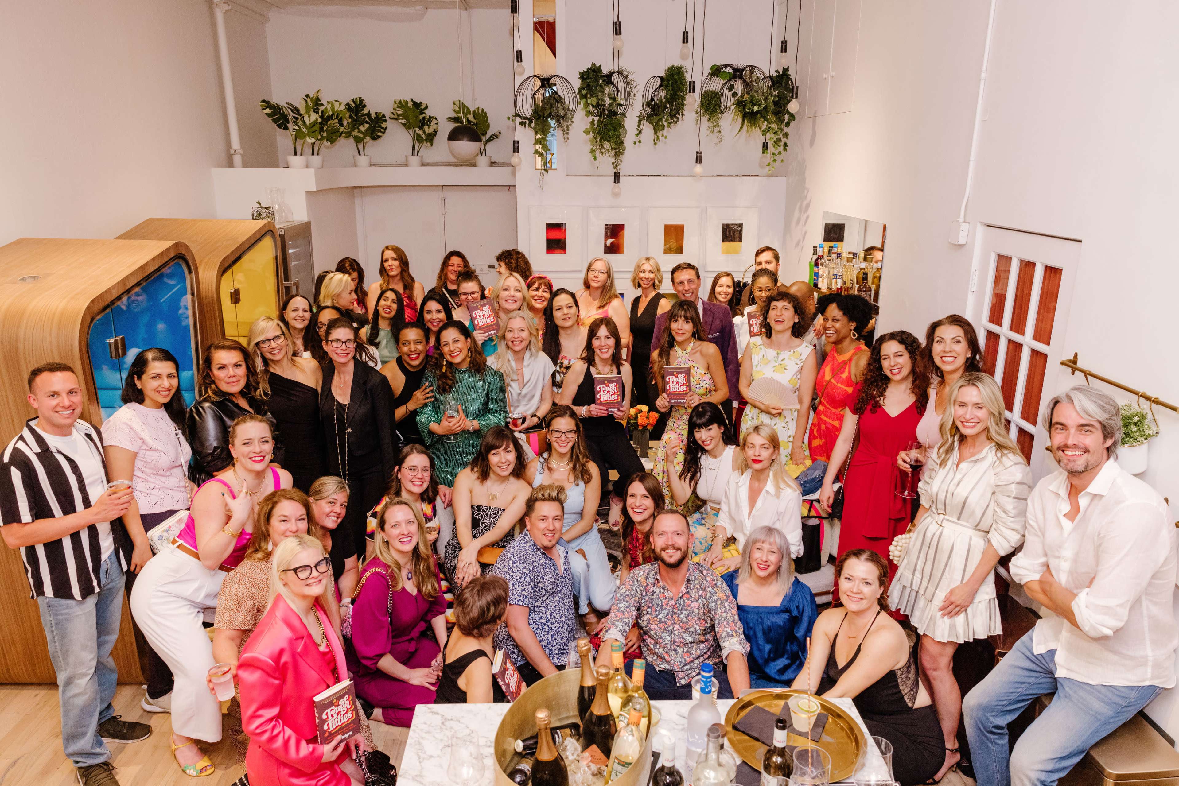 A large group of people, both men and women, gathers together in a bright, modern space featuring plants and a bar setup for a celebration or event.