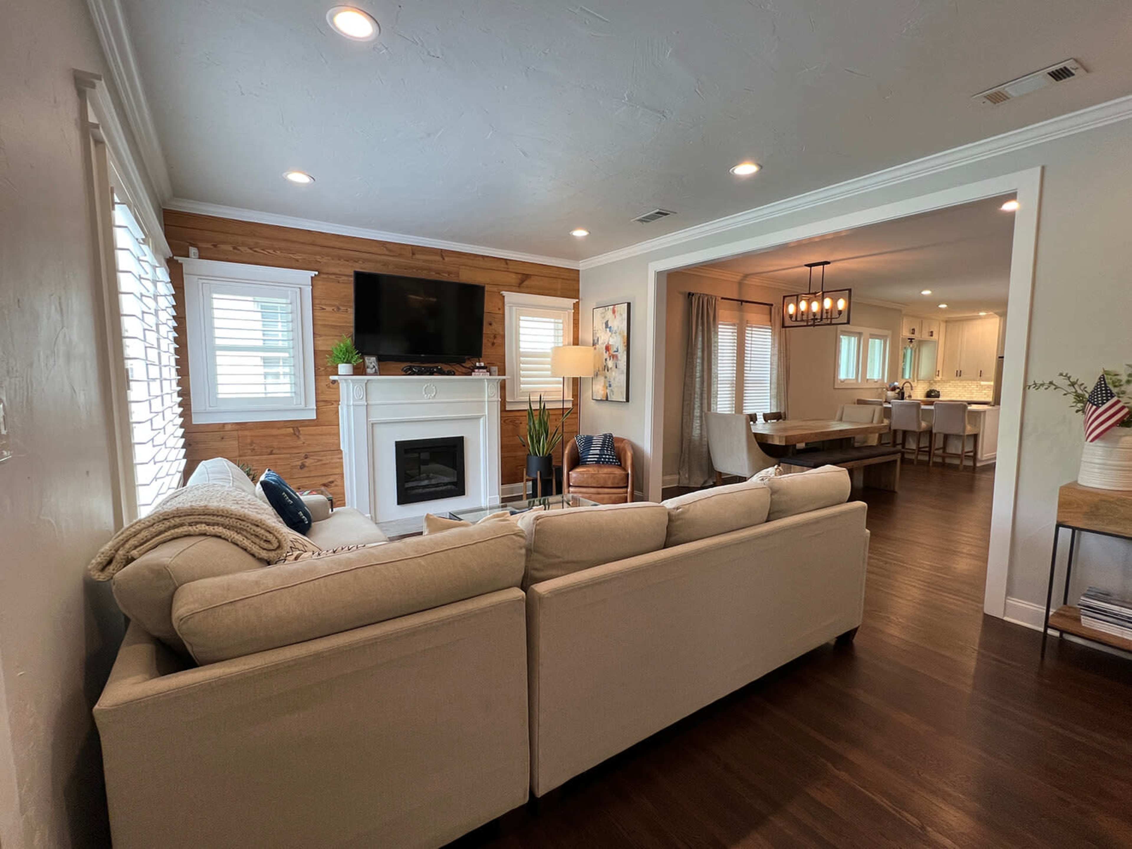 A beige sofa is positioned in a living room that features a fireplace and a wall-mounted TV, with a view into a dining area and kitchen beyond.