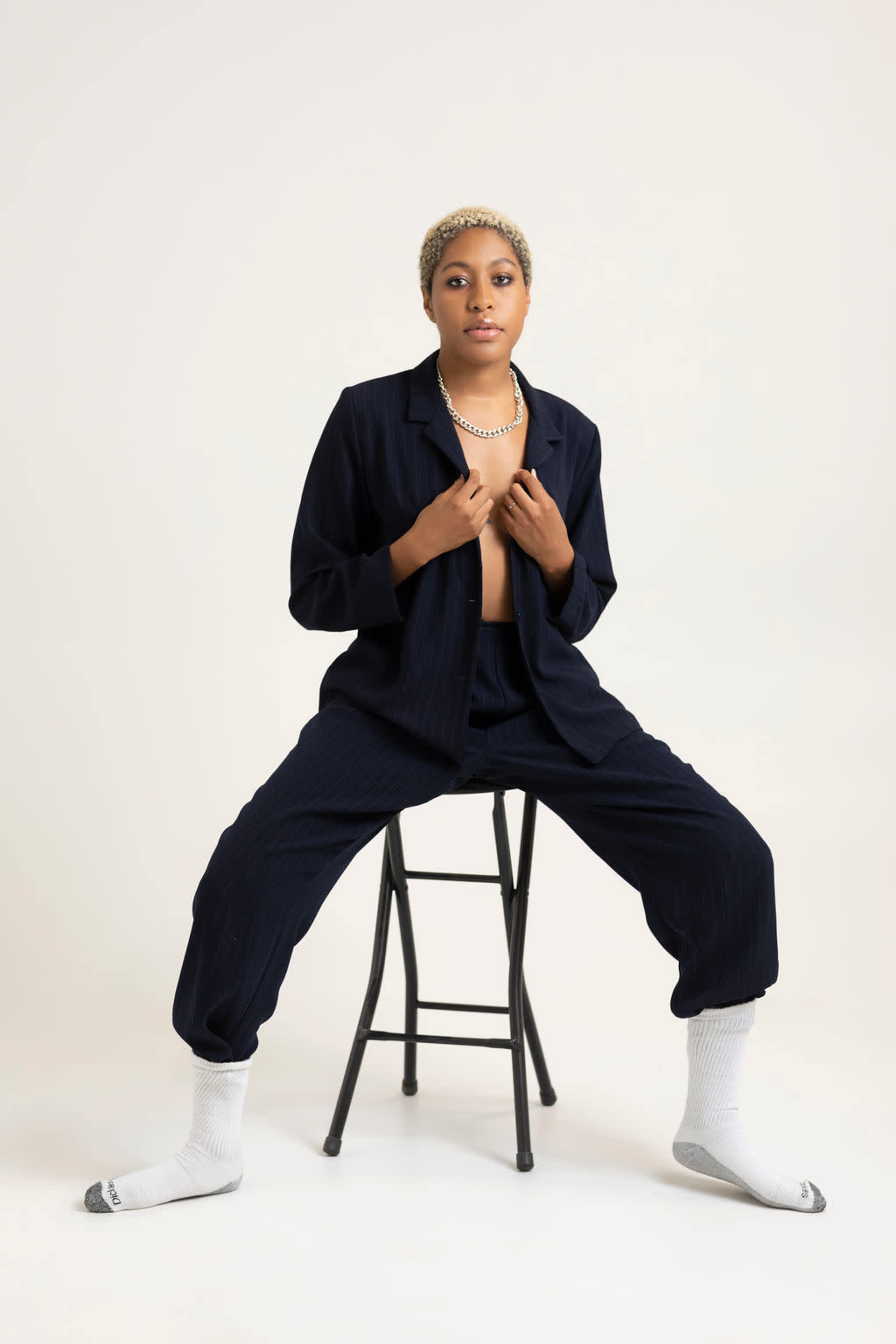 A person sits on a black stool, wearing a dark outfit, with their hands near their chest, against a plain white background.