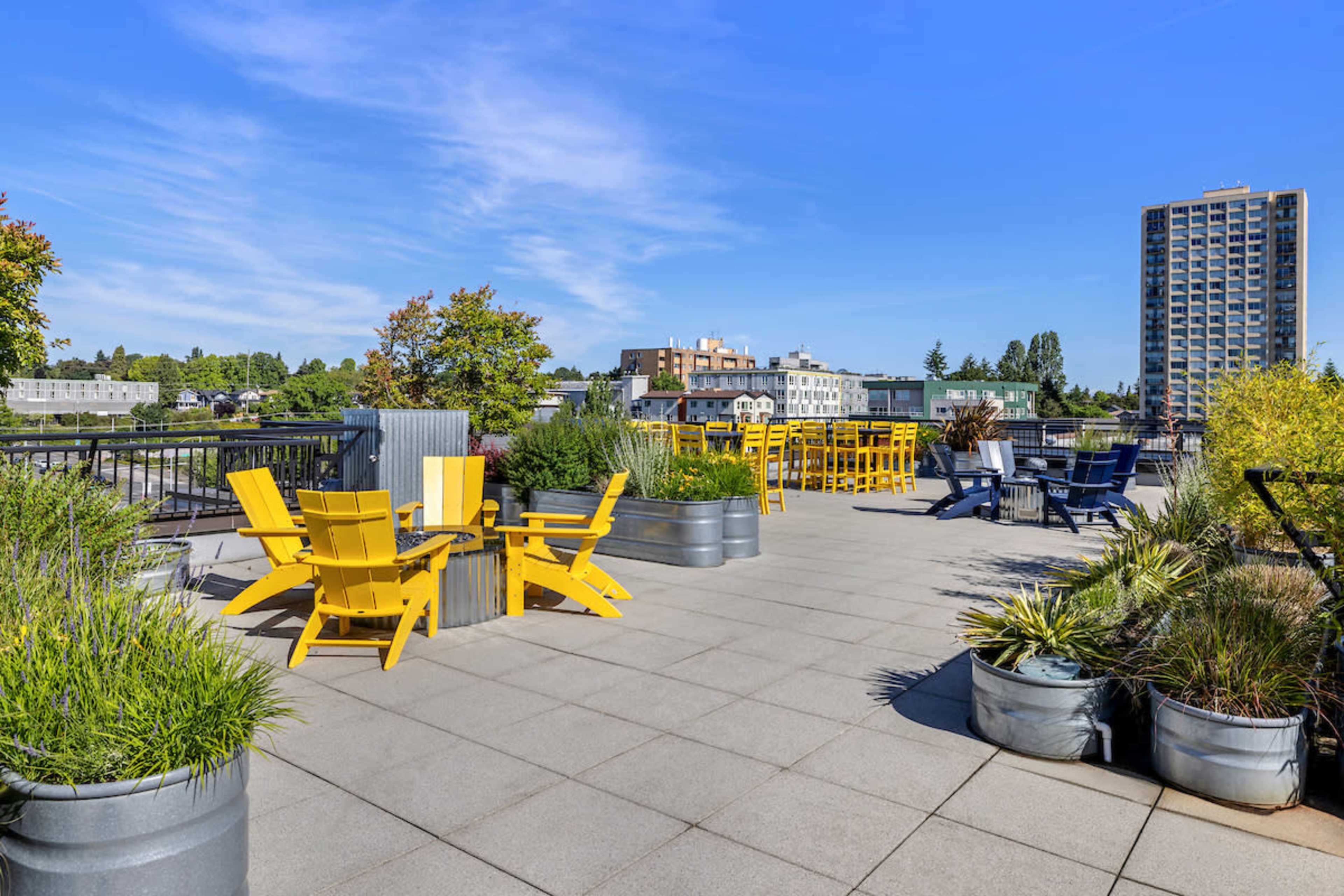 The rooftop terrace features yellow picnic tables and chairs surrounded by green landscaping and planters, with a city skyline in the background.