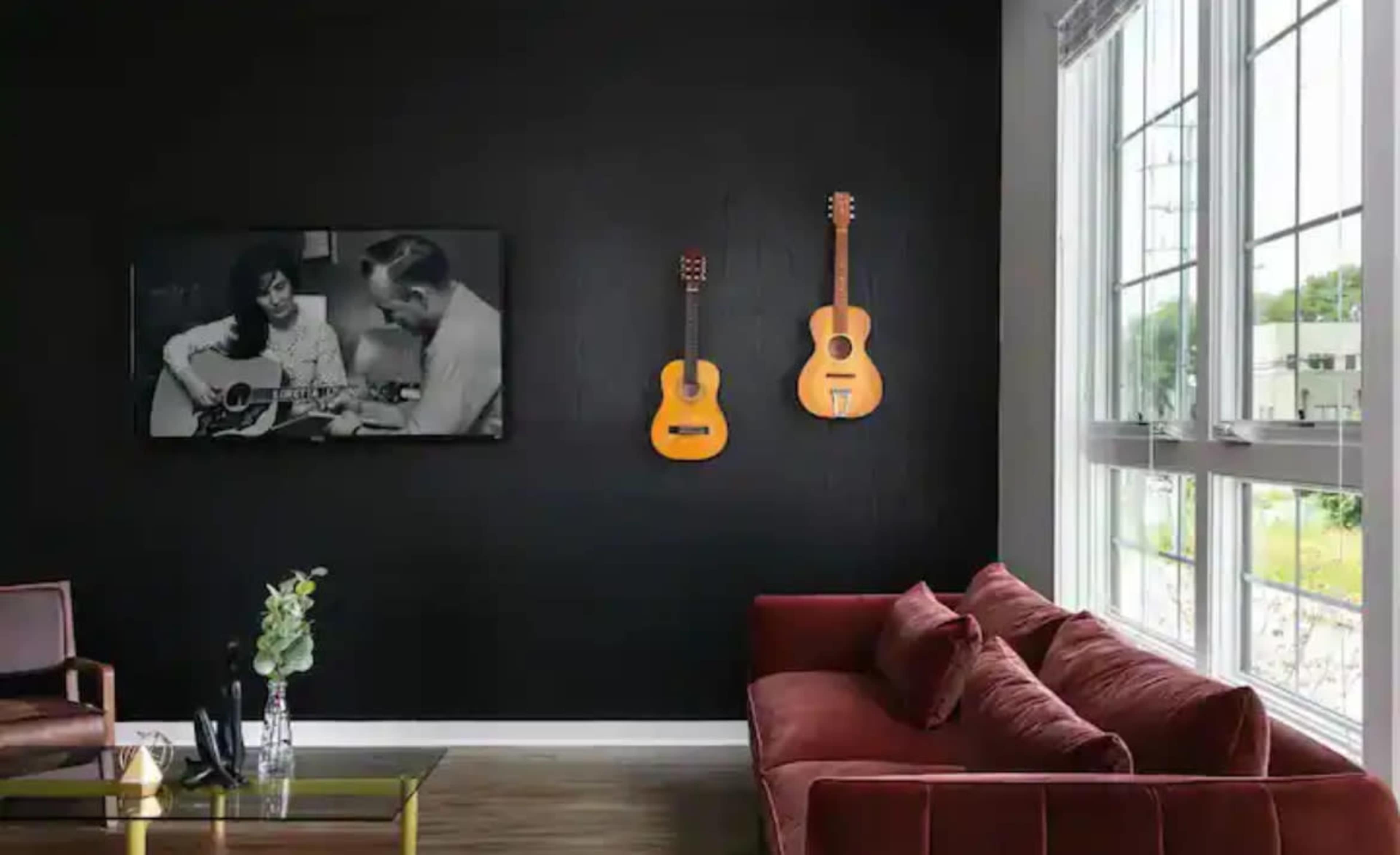 A black wall displays a black-and-white photo of two people playing guitar, while two acoustic guitars hang nearby above a red sofa and a glass coffee table.