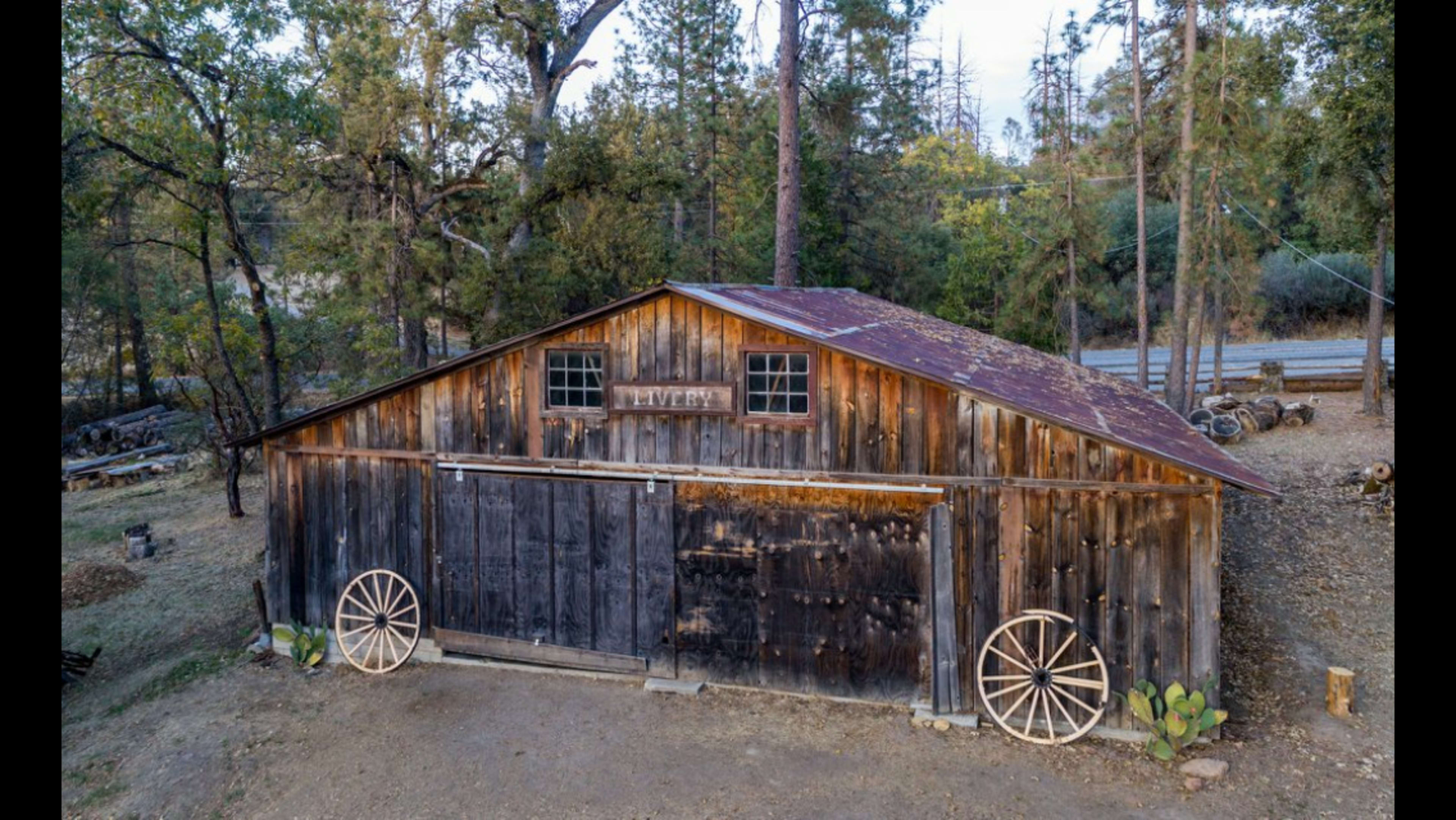 A rustic wooden barn with a red metal roof, flanked by two wooden wagon wheels and surrounded by trees.