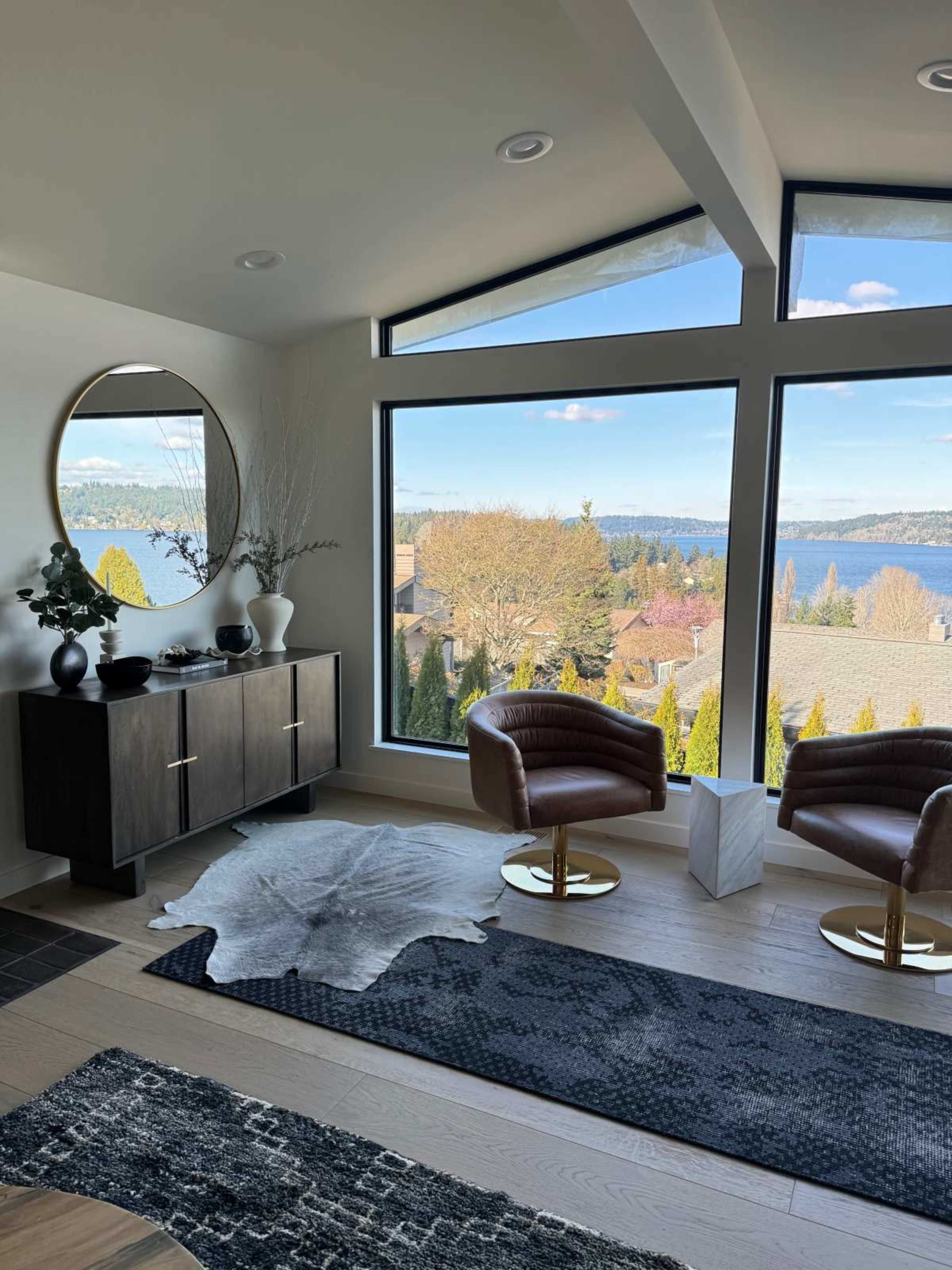 The image shows a stylish living room with two brown chairs, a modern cabinet, and large windows overlooking a scenic view of trees and water.
