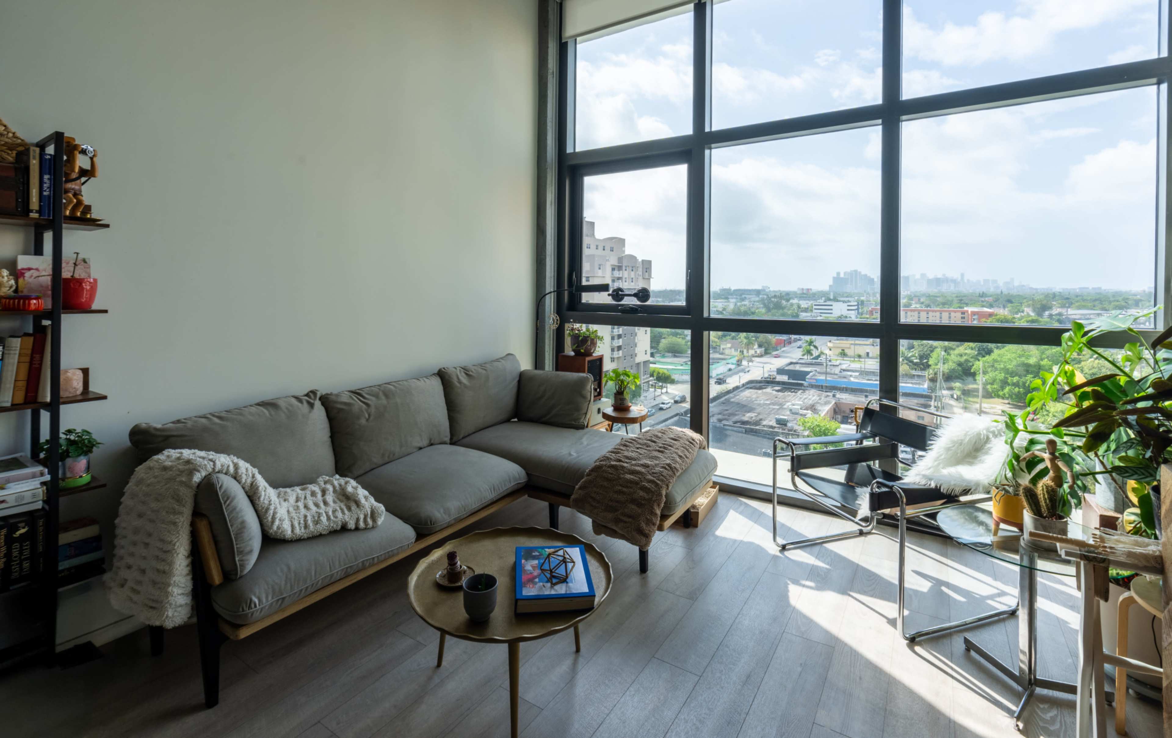 The image shows a modern living room with a gray sectional sofa, a small round coffee table, and large windows overlooking a cityscape.
