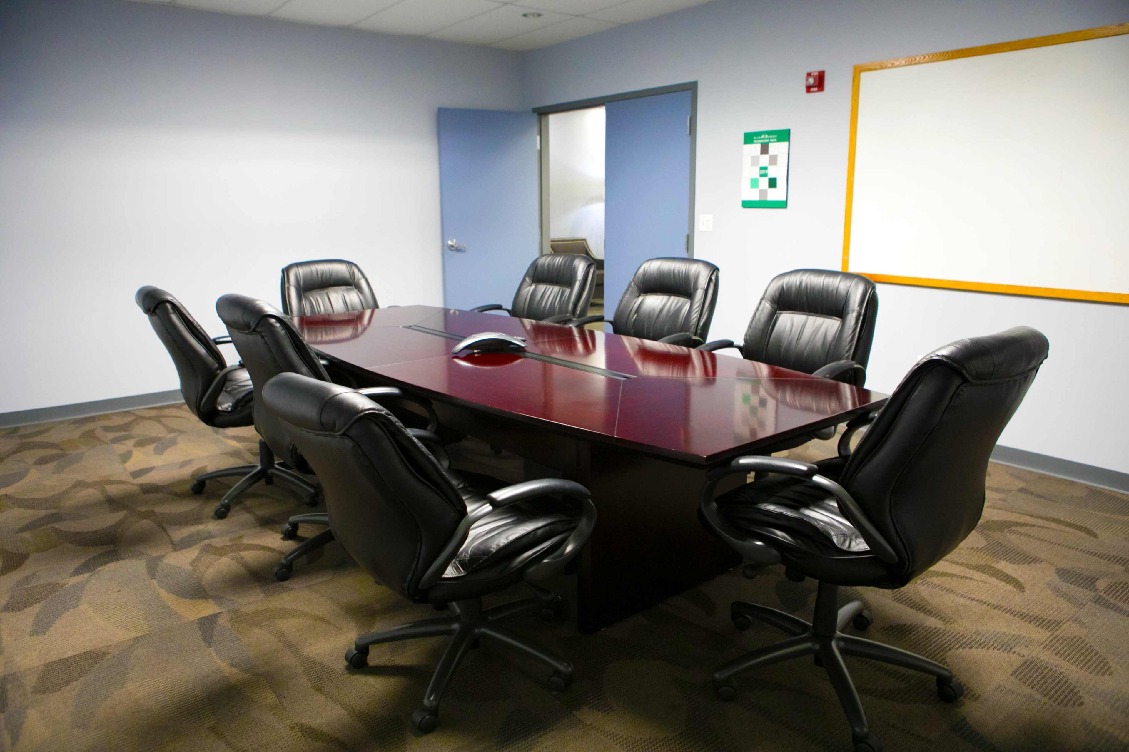 A conference room features a large wooden table surrounded by six black ergonomic chairs, with a whiteboard and emergency exit sign on the walls.