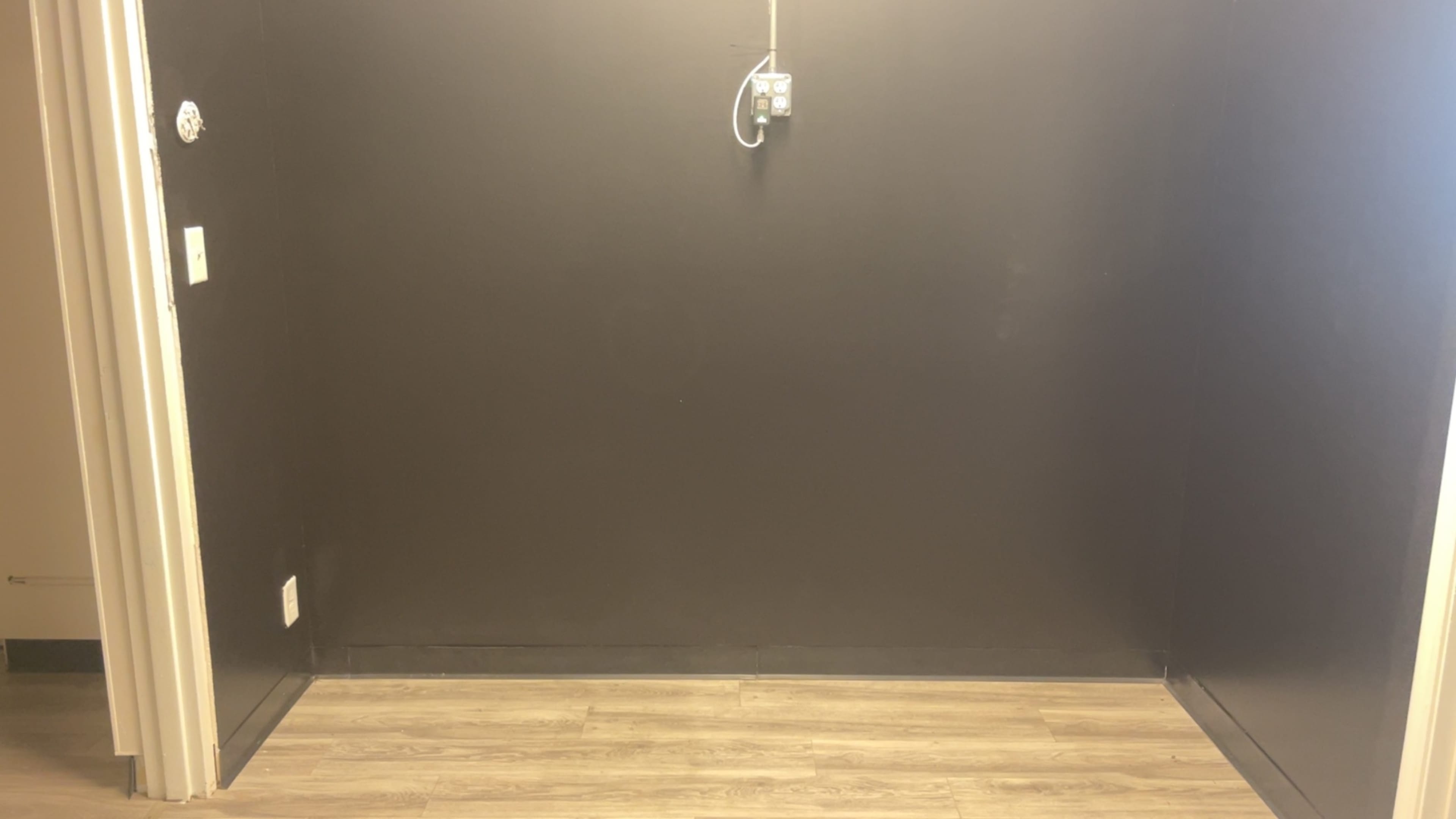 The image shows a bare room with a dark wall, hardwood flooring, and a single electrical outlet on the left.