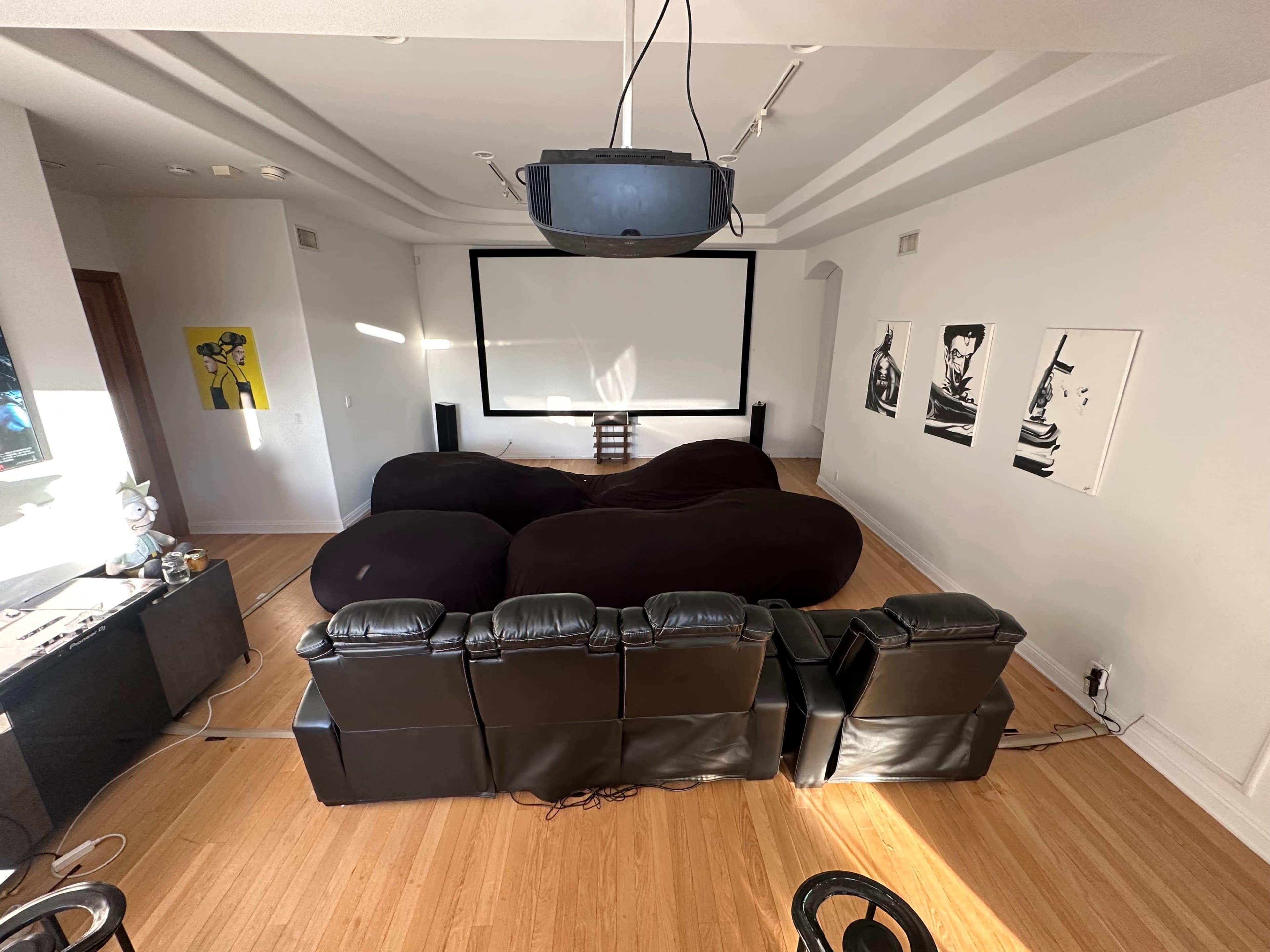 A home theater room features a large, dark sectional sofa facing a projection screen, with a row of black reclining chairs behind it and artwork on the walls.