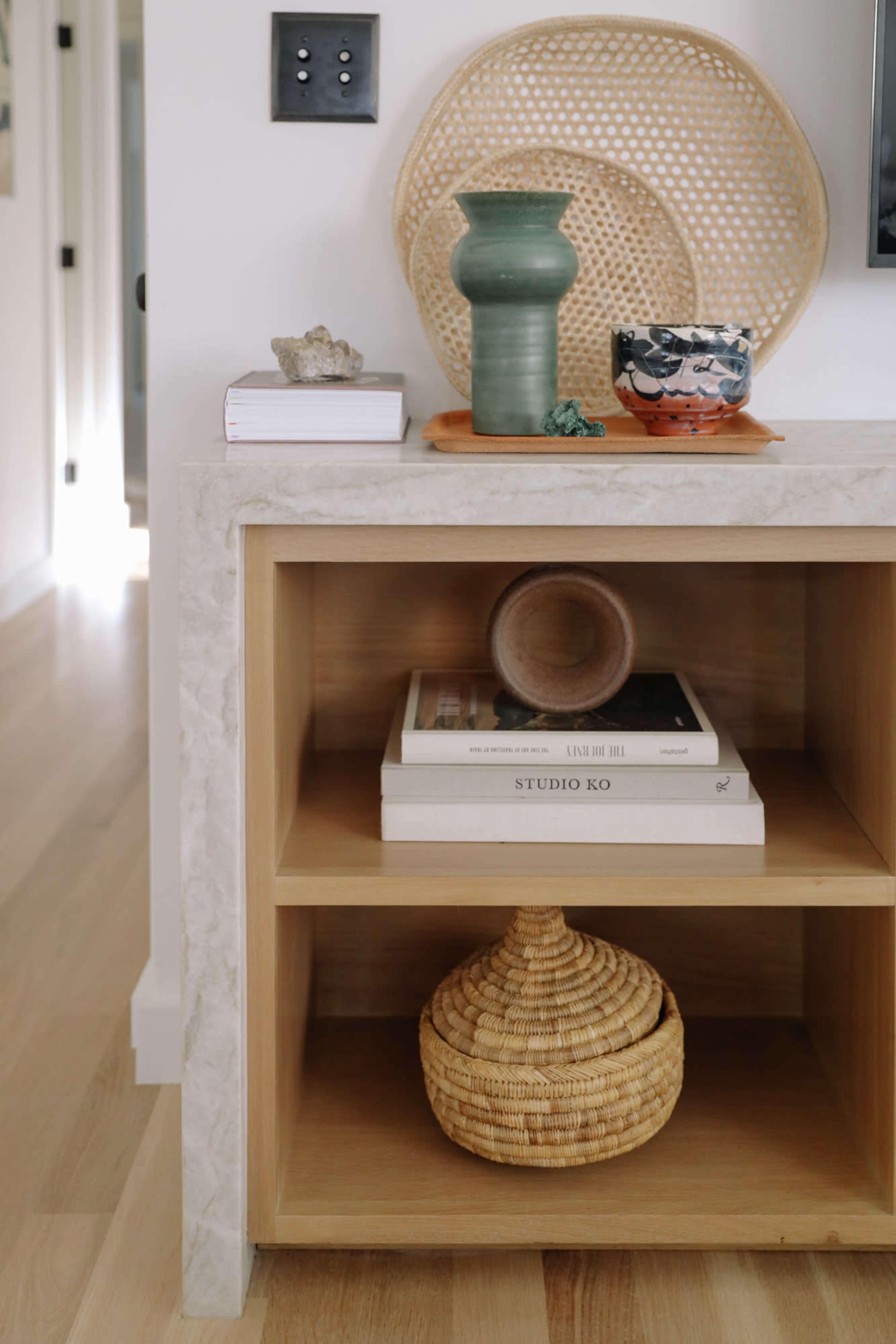 A wooden console table displays decorative items, including a green vase, a woven plate, and stacked books, with a round wicker basket on the bottom shelf.