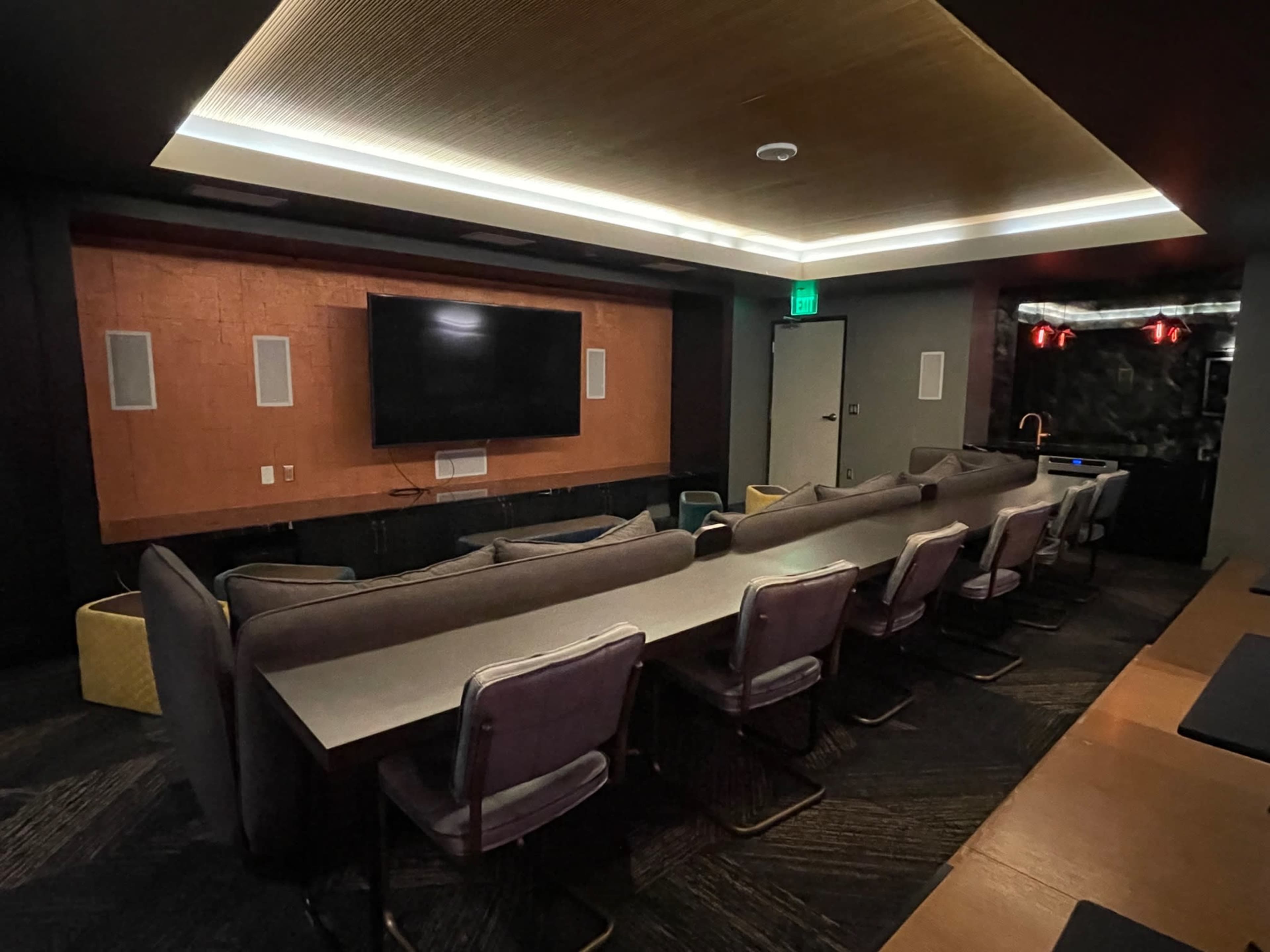 A modern, dimly lit room with a long table surrounded by chairs, a large wall-mounted television, and a textured wall panel behind the seating area.