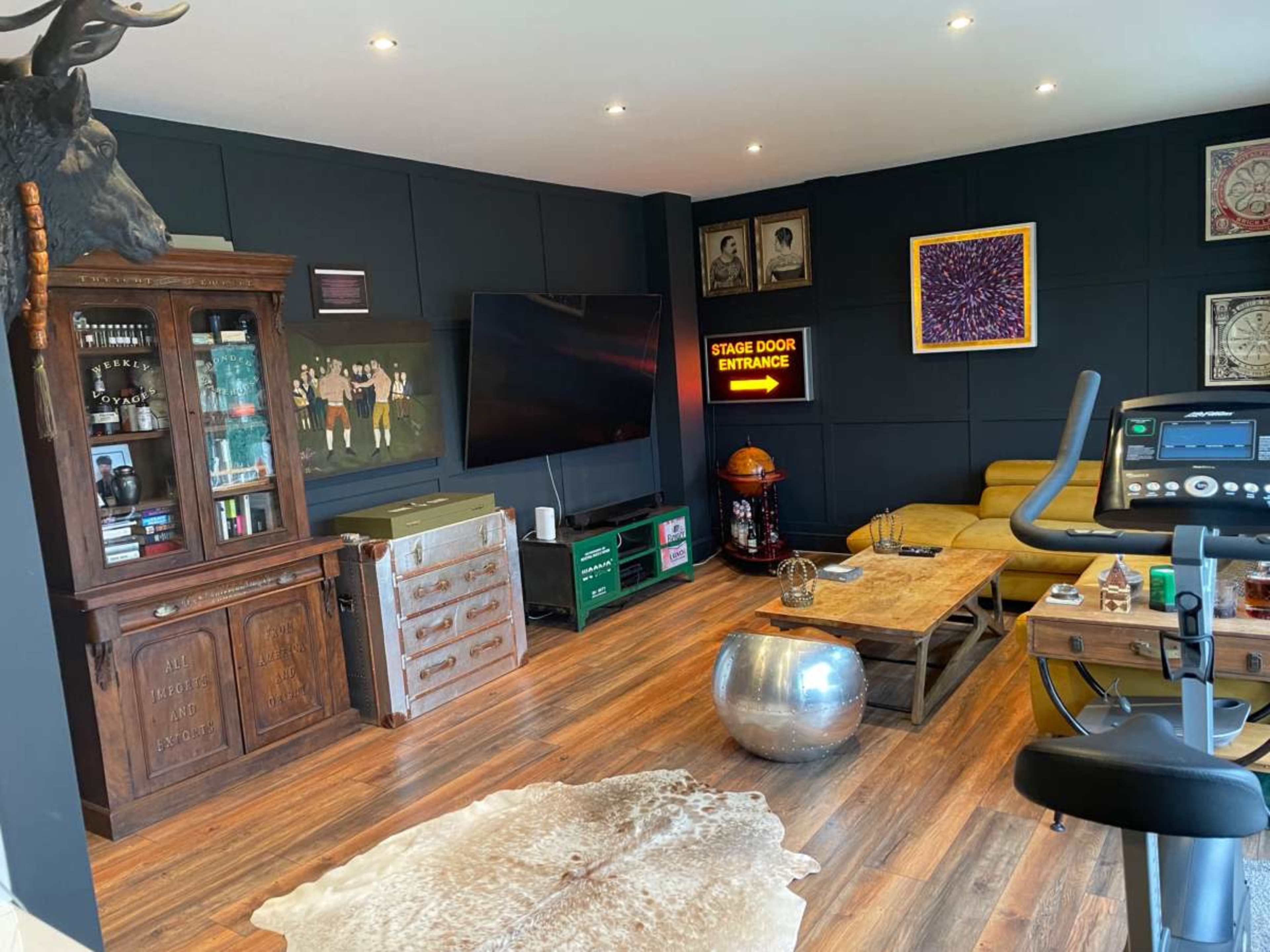 The room features a modern living space with a fitness bike, a wooden cabinet, a leather sofa, and a large television on a black wall.
