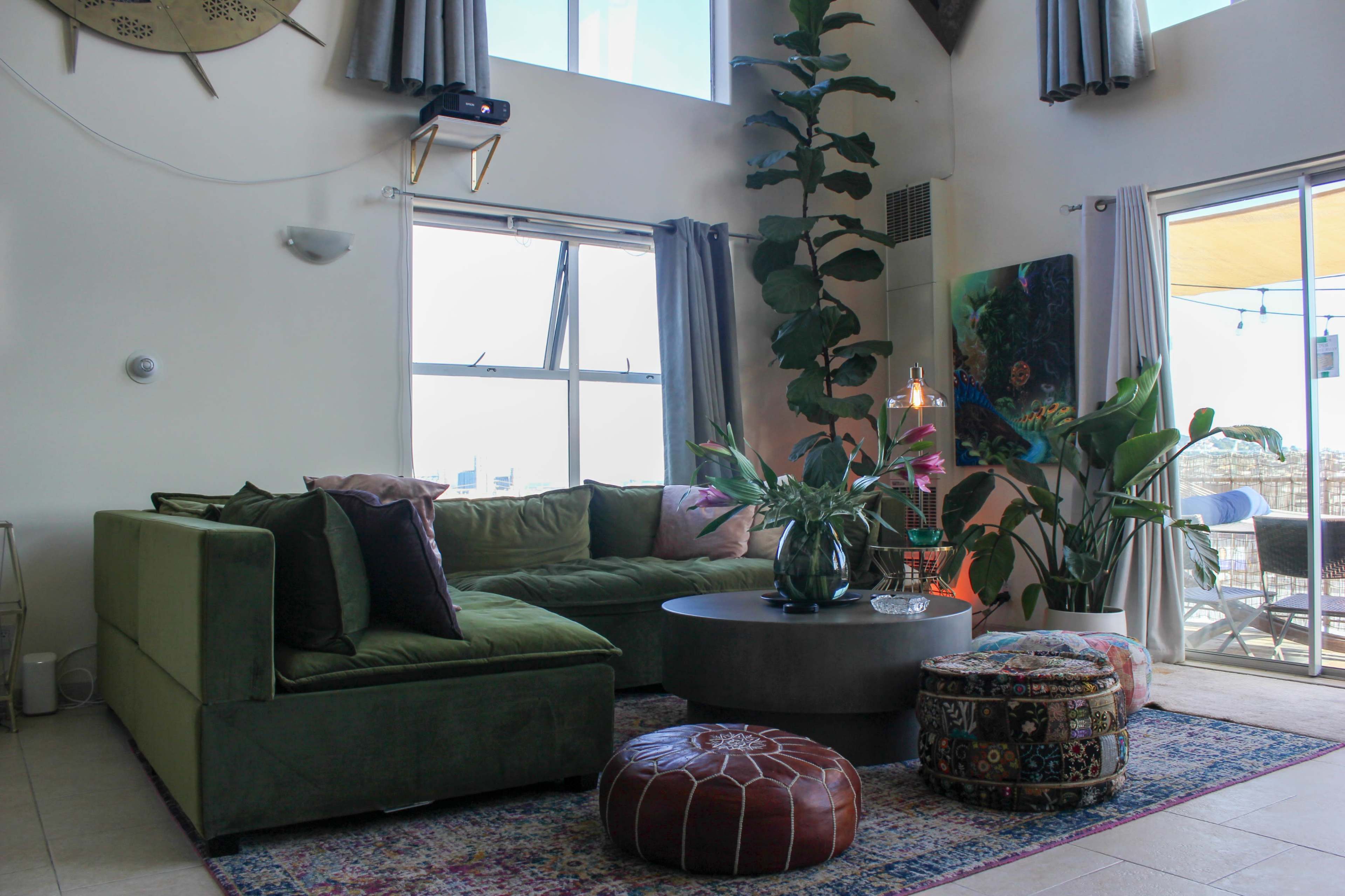 The image shows a modern living room featuring a green sectional sofa, a round coffee table, and a tall indoor plant, with large windows providing natural light.