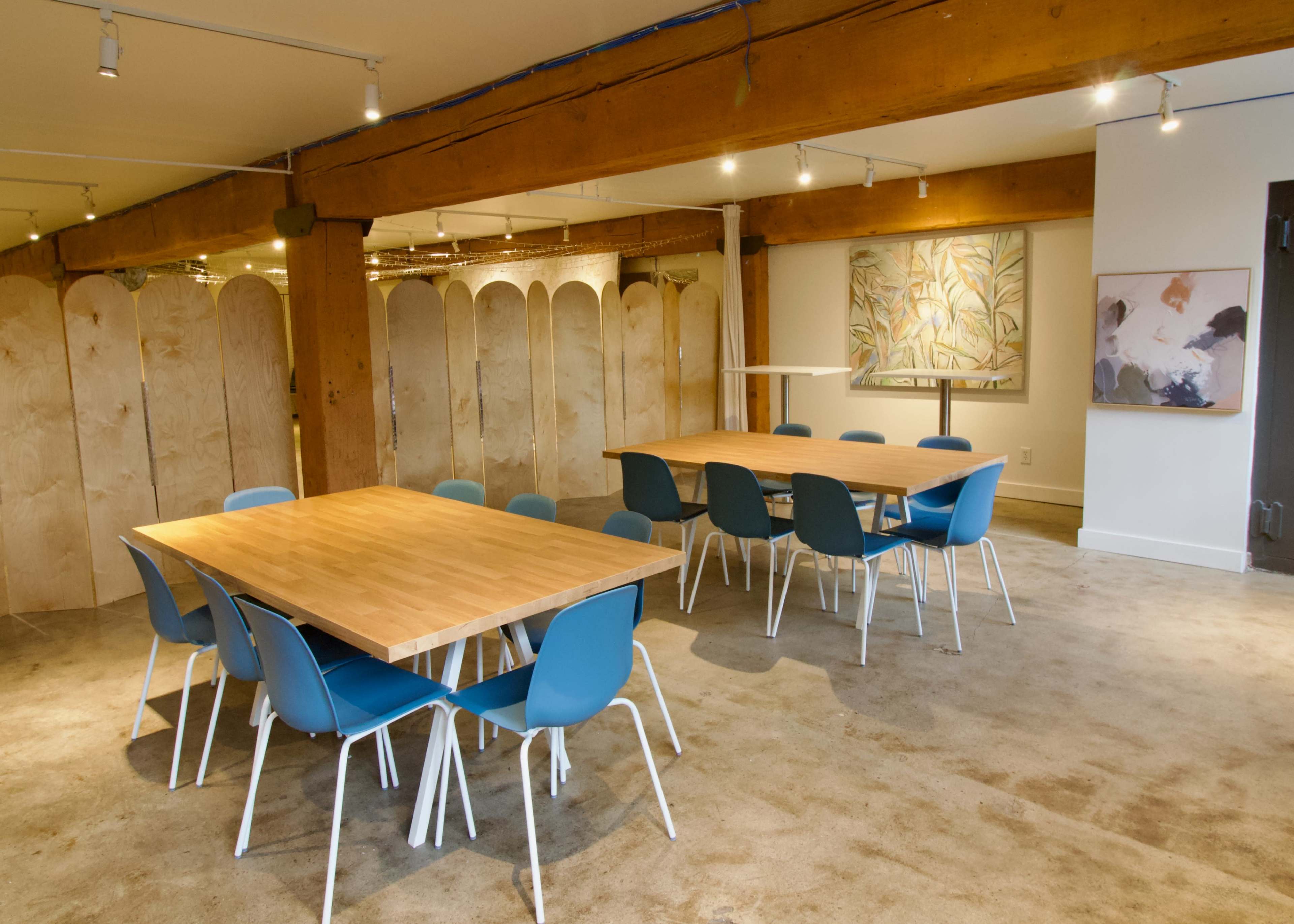 The image shows a modern meeting room featuring two wooden tables surrounded by blue chairs, with a wooden beam overhead and decorative artwork on the walls.