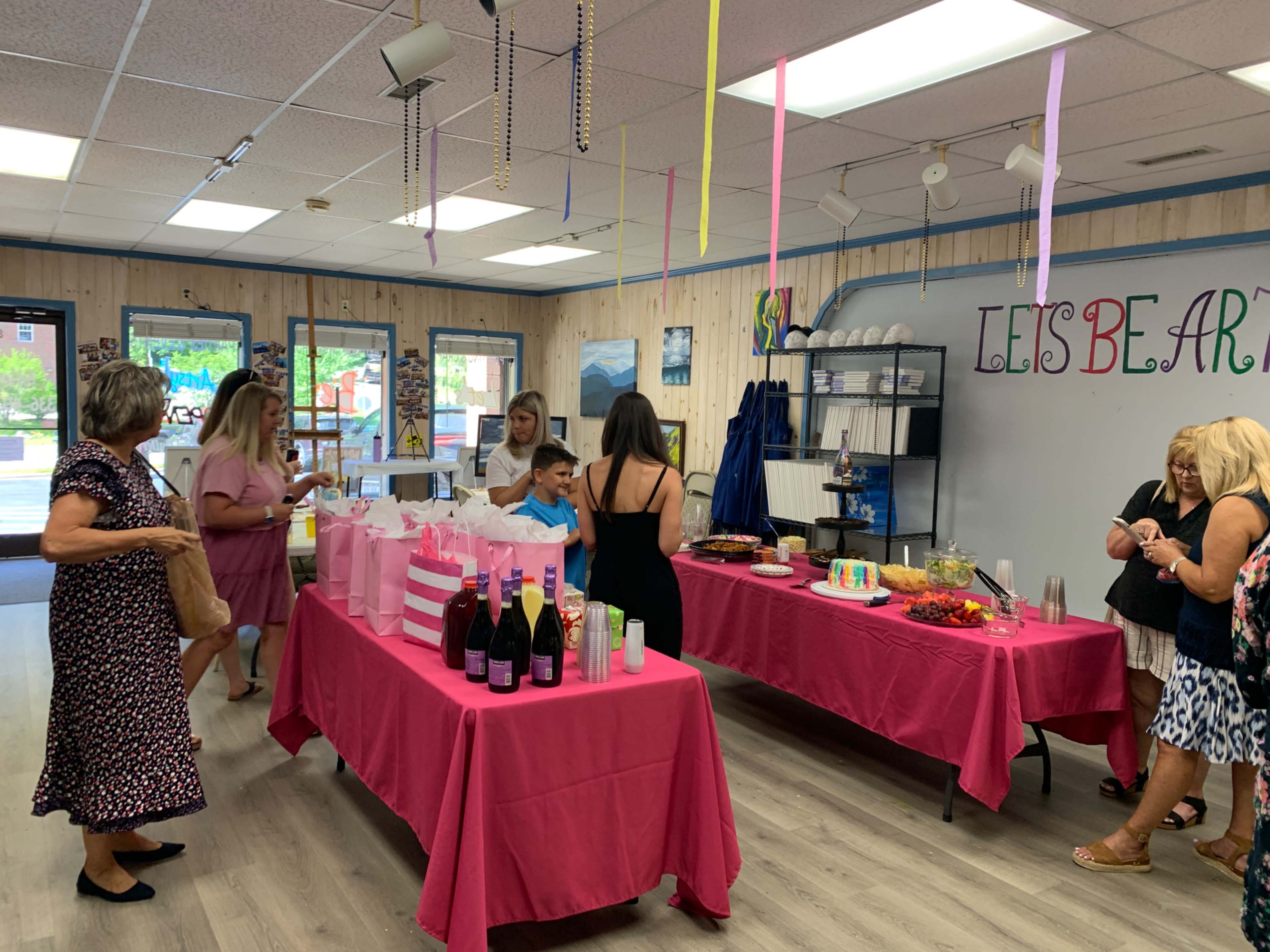 A celebration venue with guests mingling around pink tables adorned with food and drinks.
