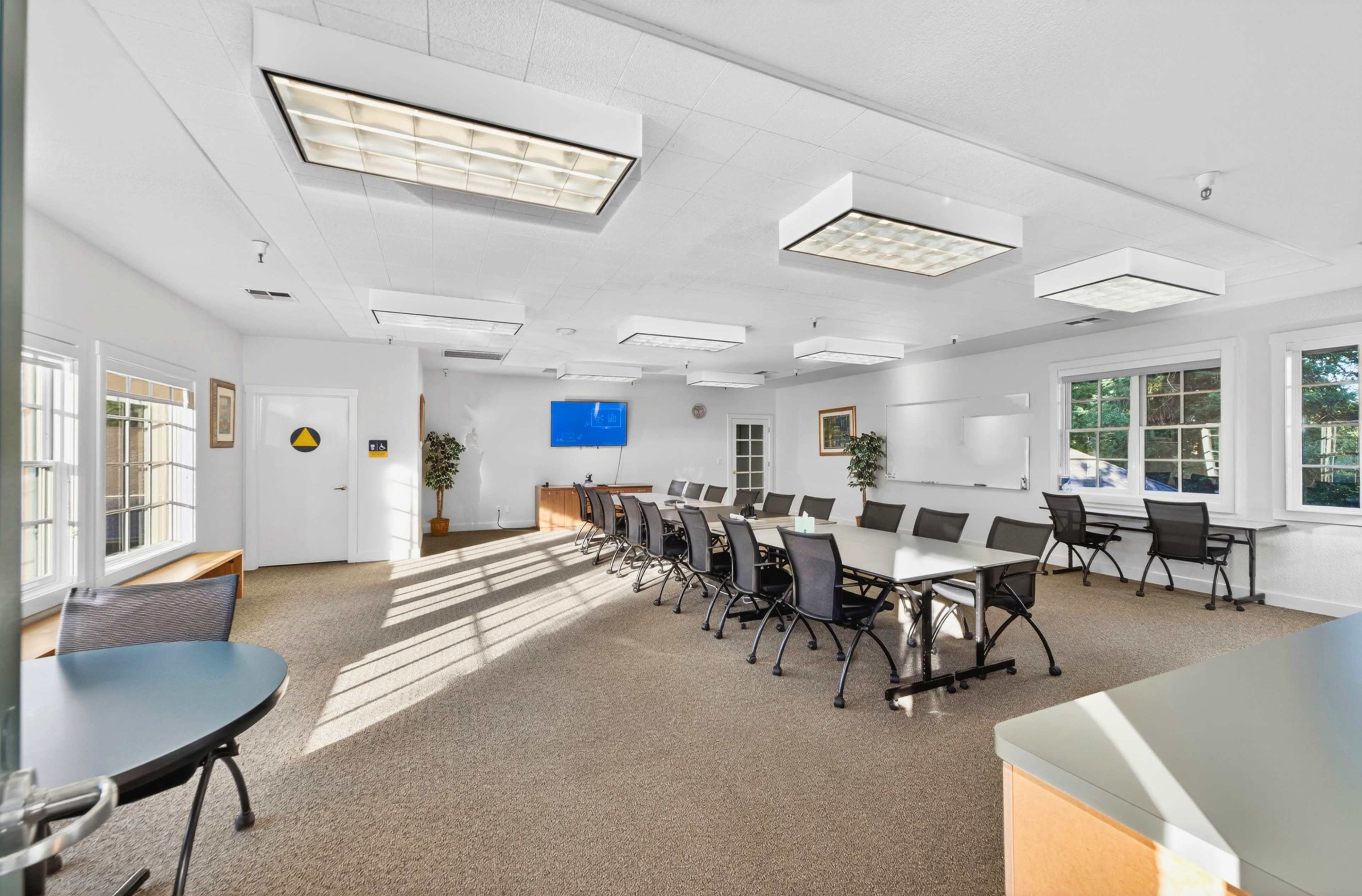 A bright conference room features a long table surrounded by black chairs, with large windows allowing natural light to illuminate the space.
