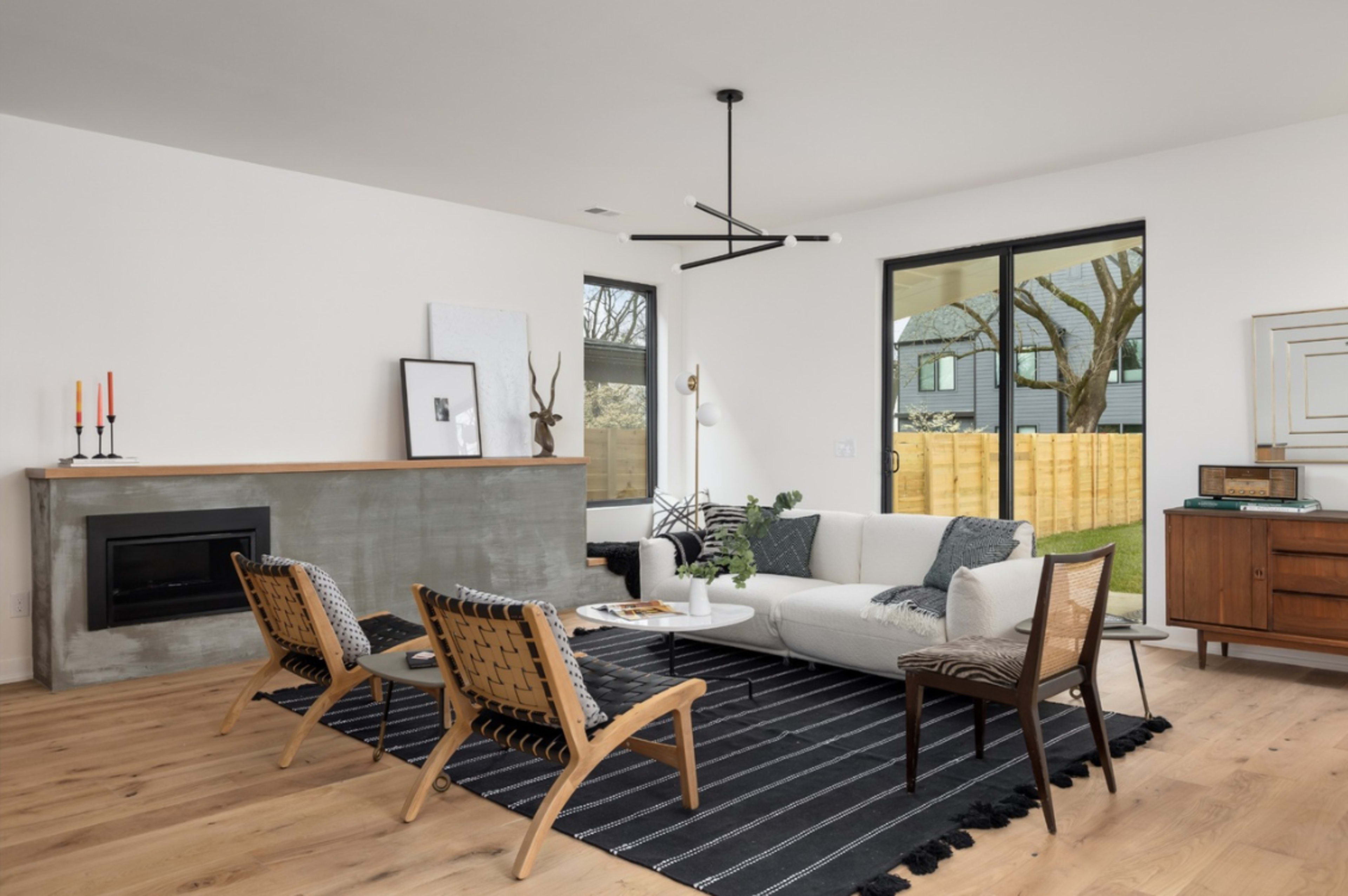 A modern living room features a concrete fireplace, a white sofa, and wooden chairs arranged on a black and white striped rug.