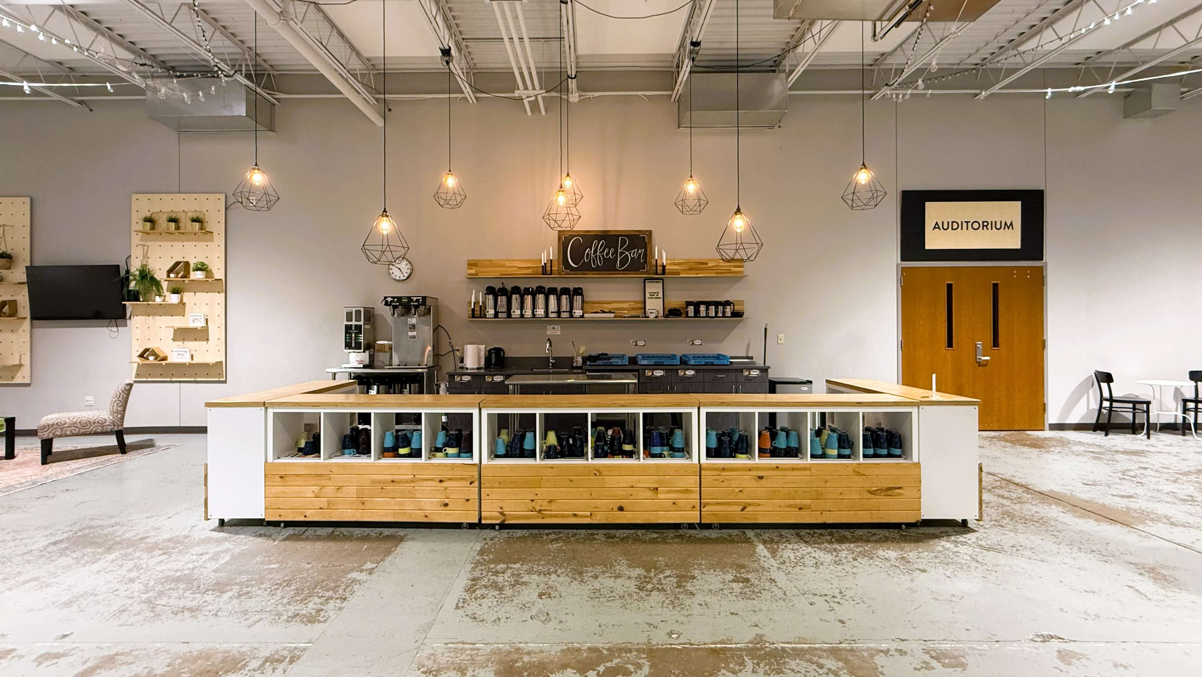 The image shows a spacious coffee bar with an elevated wooden counter, shelves displaying coffee supplies, and pendant lighting, adjacent to an auditorium entrance.