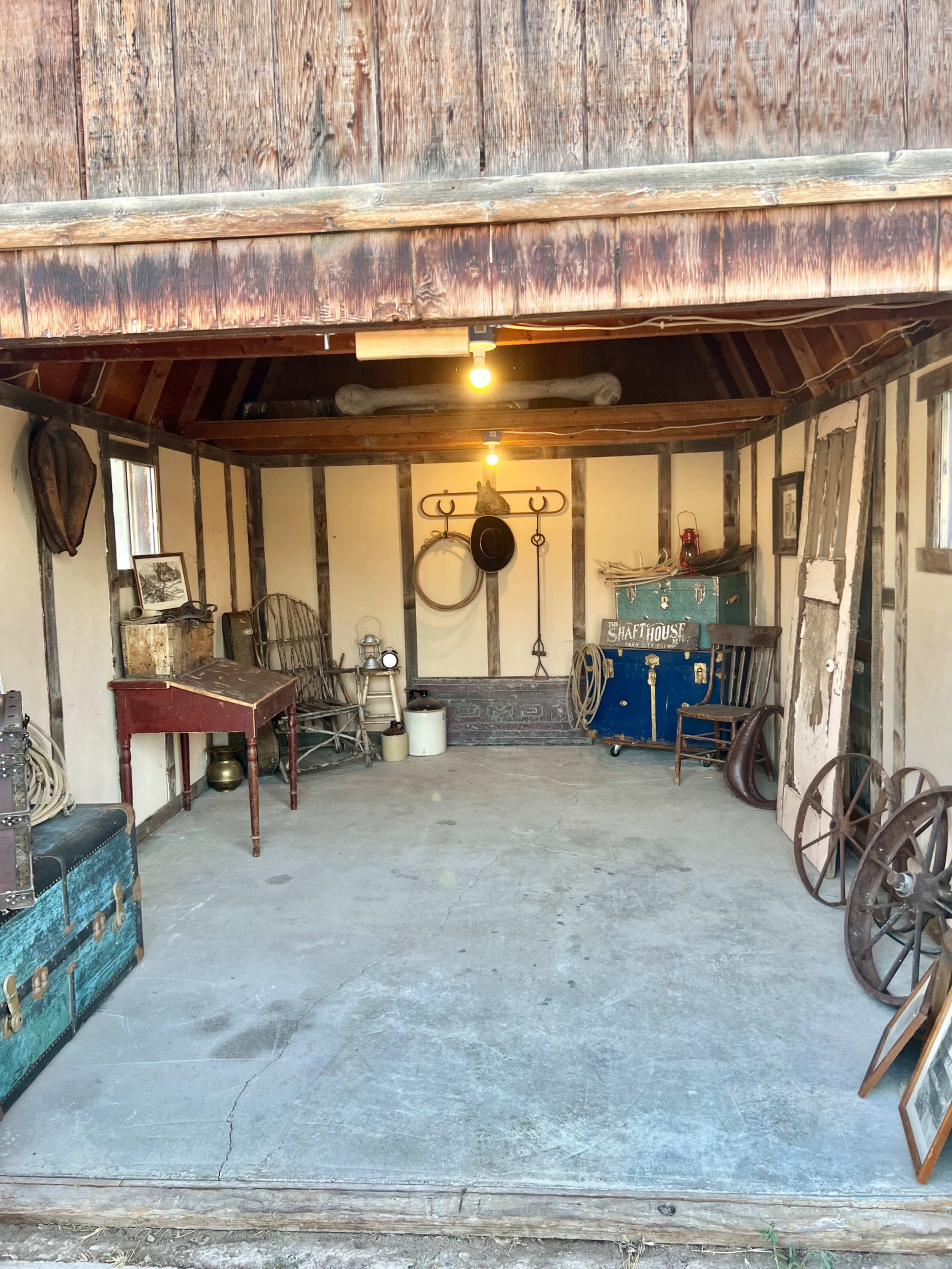 The image shows a rustic, empty garage with wooden beams, old furniture, and various vintage items, including wheels and a trunk.