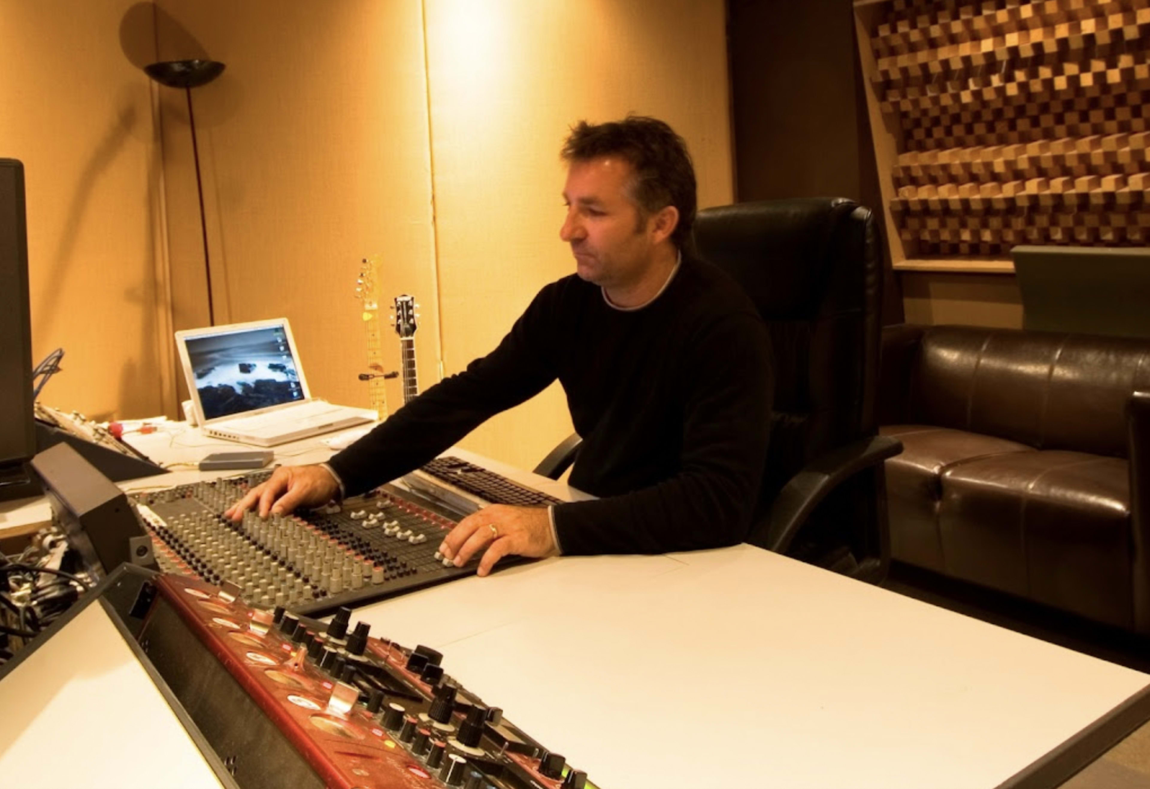 A man operates a sound mixing console in a recording studio while a computer and musical instruments are visible nearby.