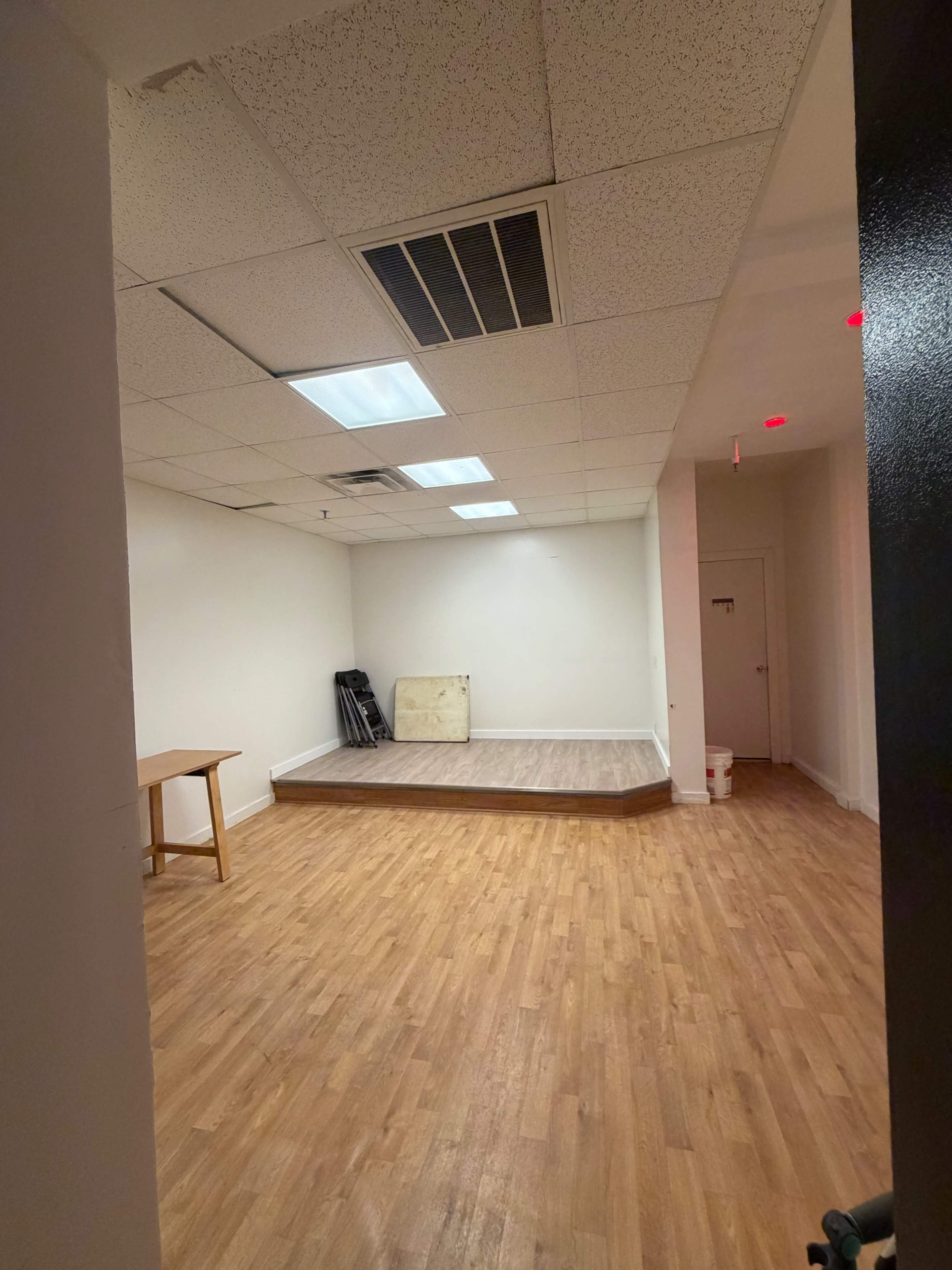 The image shows an empty interior room with a wooden floor, a raised stage area, and a table against one wall.
