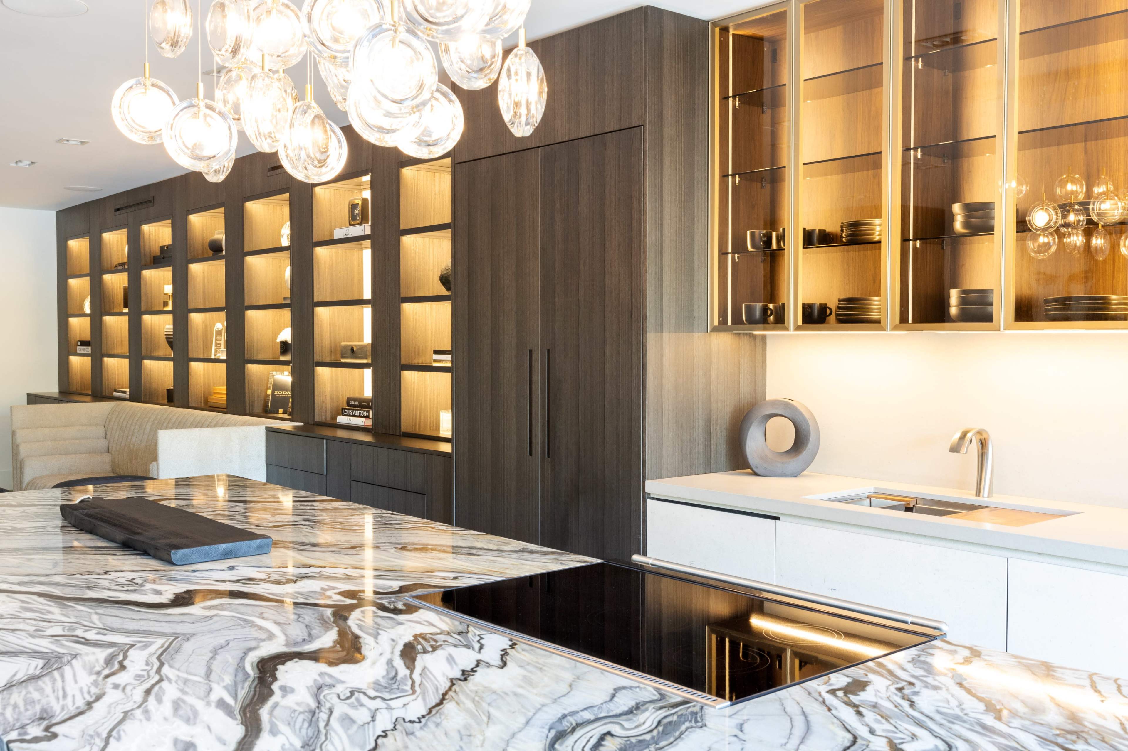 A modern kitchen features a glossy marble island, sleek cabinetry, and illuminated shelving showcasing dinnerware.