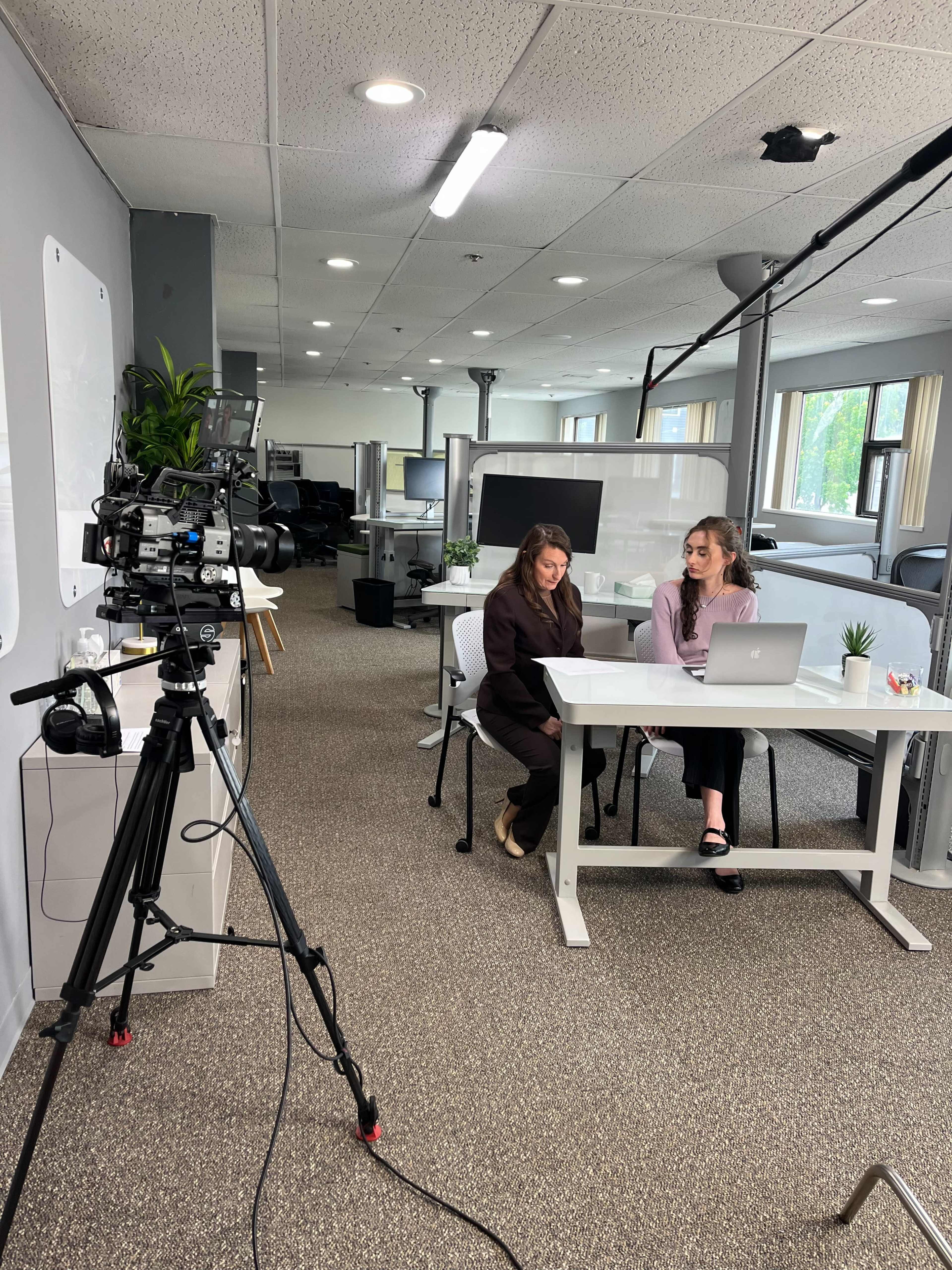 A camera is set up in a modern office space where two women are seated at a table with a laptop, discussing something while surrounded by office furniture and plants.