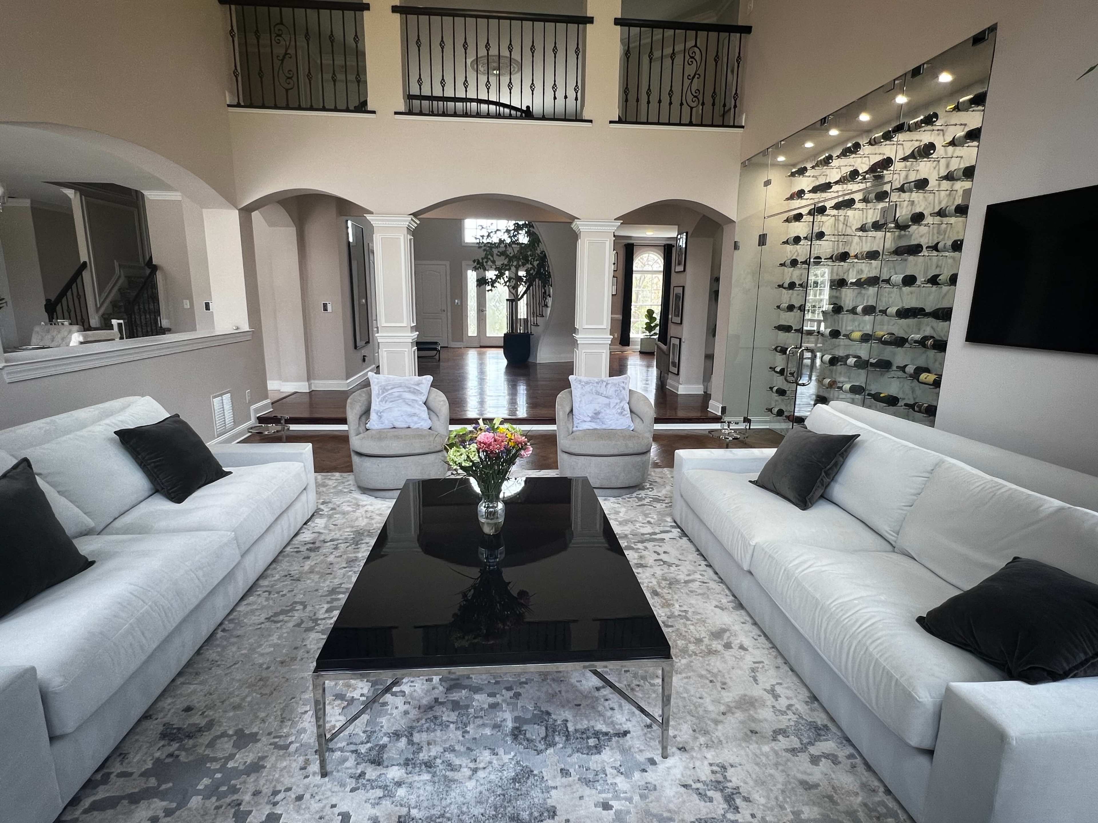 The spacious living room features two light-colored sofas and two armchairs arranged around a sleek black coffee table, with a wine display wall visible in the background.