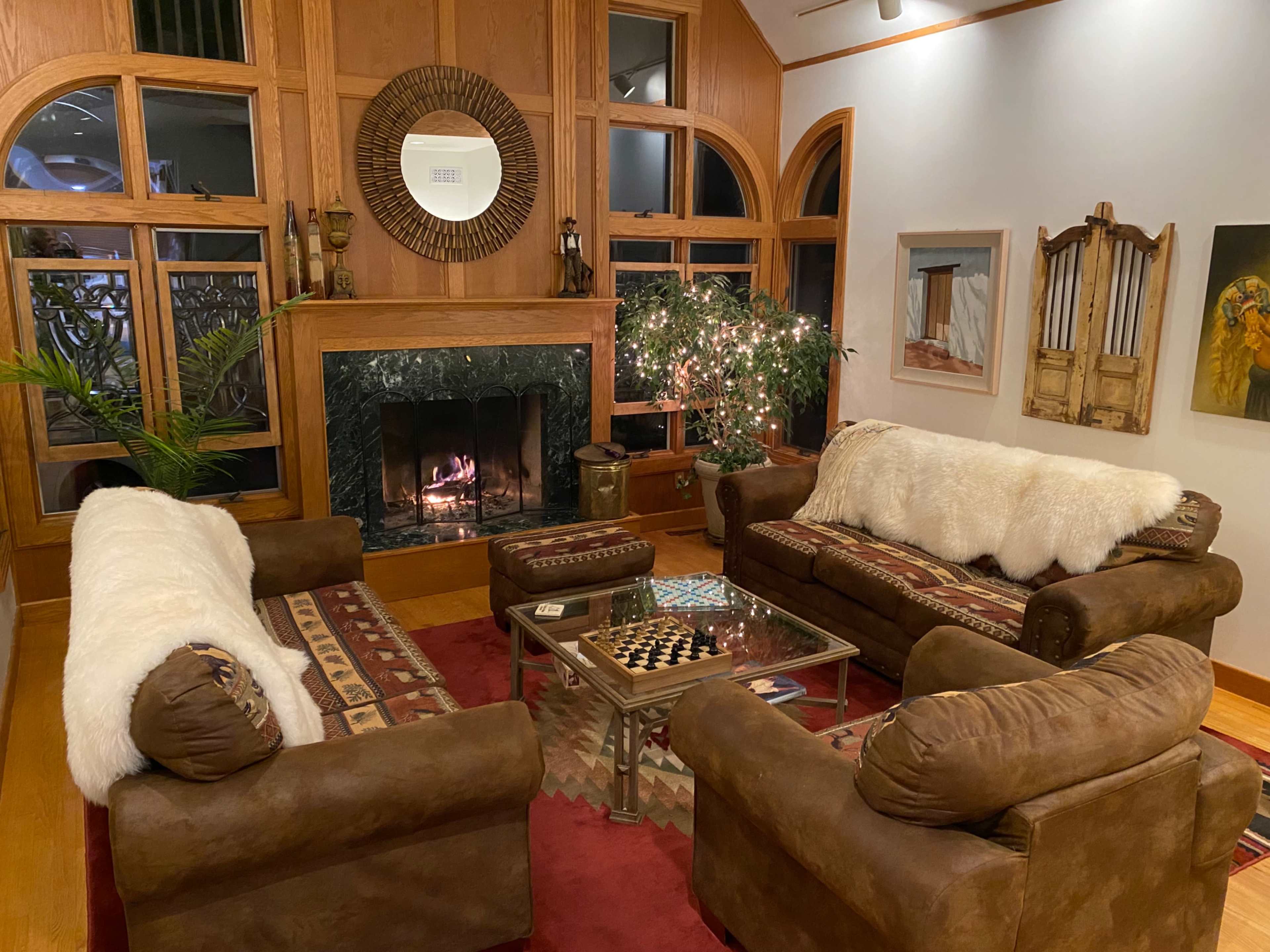 The scene features a cozy living room with two brown sofas, a glass coffee table, a warm fireplace, and a decorative plant surrounded by various artworks on the walls.